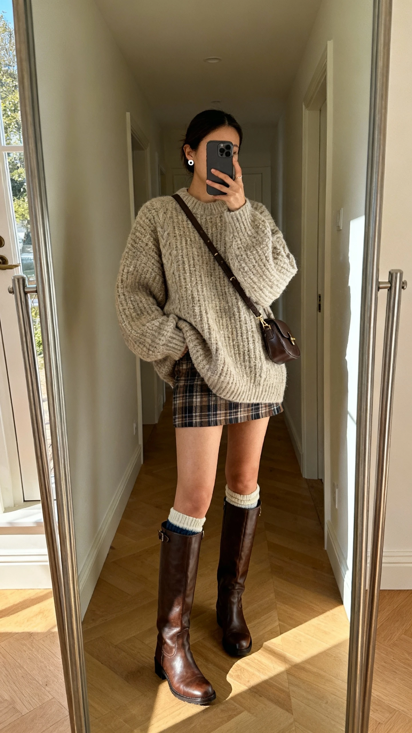 iPhone mirror selfie of a woman wearing an oversized chunky sweater, plaid mini skirt, tall knee-high leather boots, crossbody bag, cozy socks peeking, simple stud earrings, face not visible, full-length hallway mirror, soft afternoon light, natural iPhone photo.
