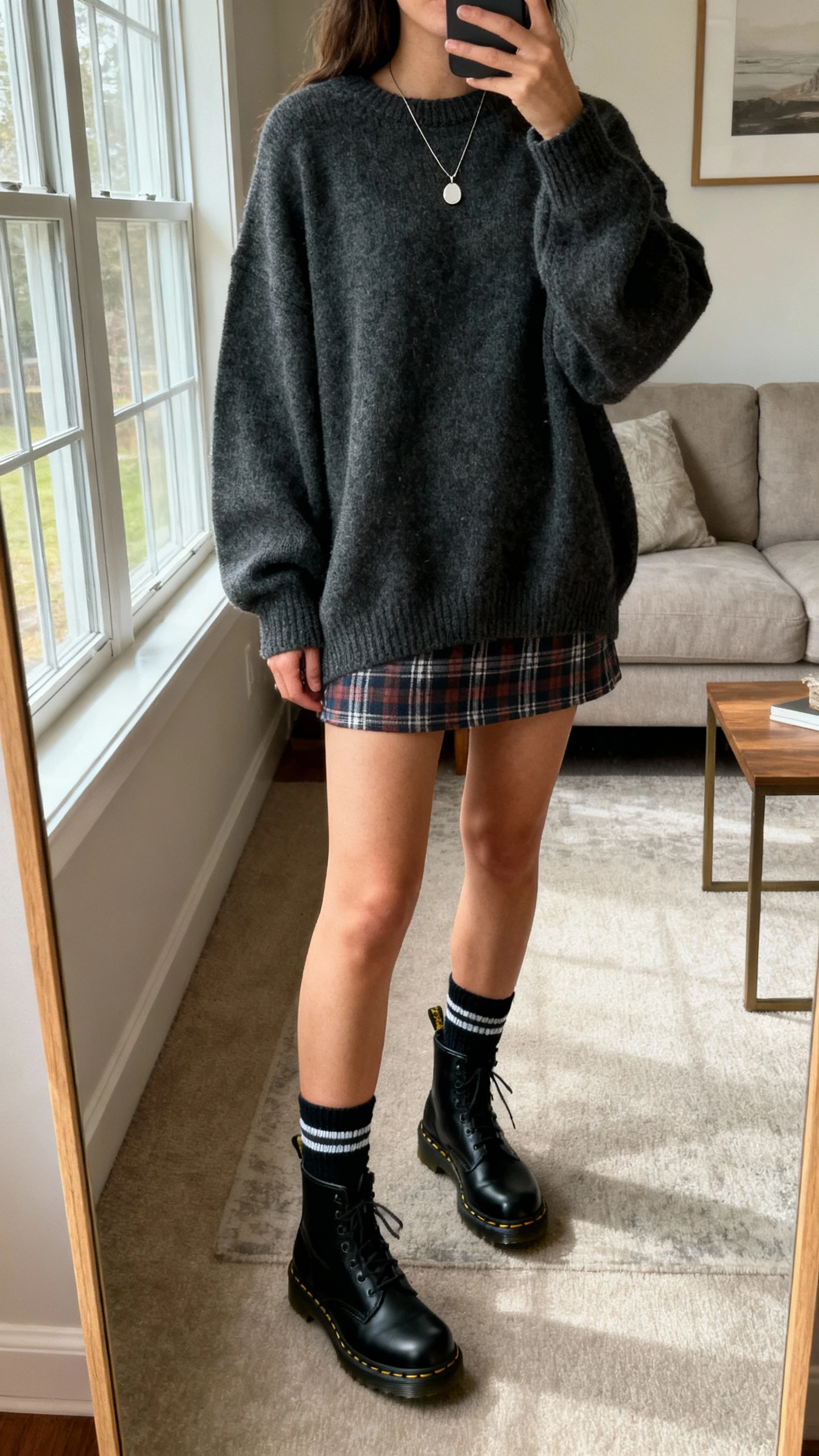 iPhone mirror selfie of a woman wearing an oversized charcoal sweater, plaid mini skirt, black Doc Martens with crew socks, simple pendant necklace, face not visible, living room full-length mirror, morning window light, natural iPhone photo quality.