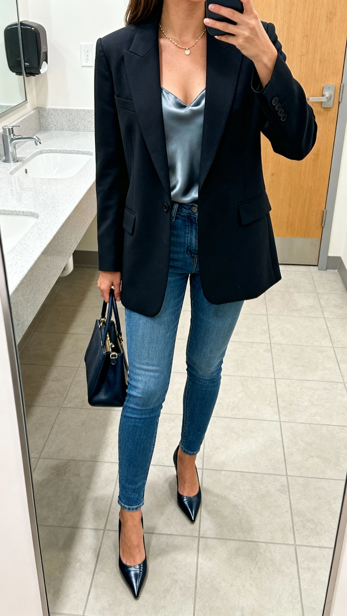 iPhone office bathroom mirror selfie of a woman in a tailored blazer over a silky camisole with slim blue jeans, pointed-toe heels, delicate necklace, structured tote, face not visible, cropped at neck, bright indoor lighting, natural iPhone quality.