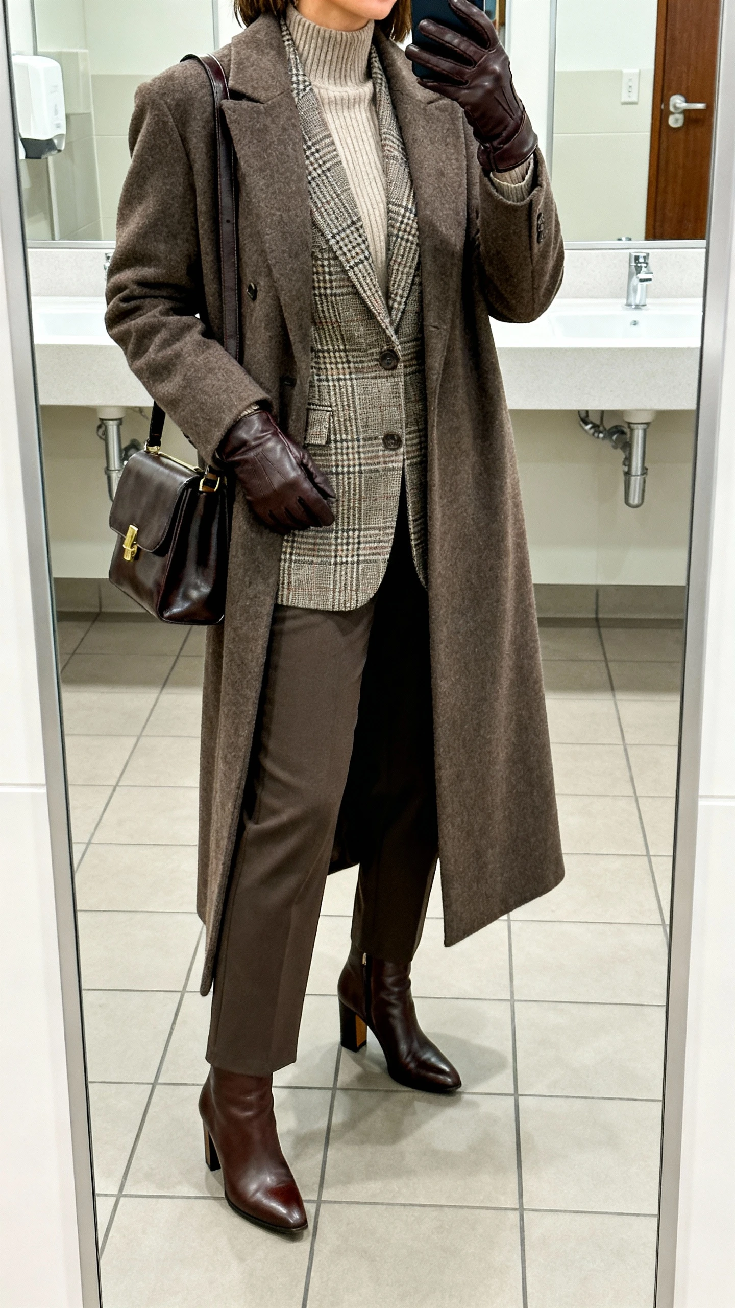 iPhone office bathroom mirror selfie of a woman layering a long ankle-grazing wool-blend maxi coat over a structured plaid blazer, fine-knit mock-neck top, tailored straight-leg trousers, leather heeled ankle boots, a polished shoulder bag and leather gloves; cropped at neck, face not visible, indoor lighting, natural iPhone photo quality.