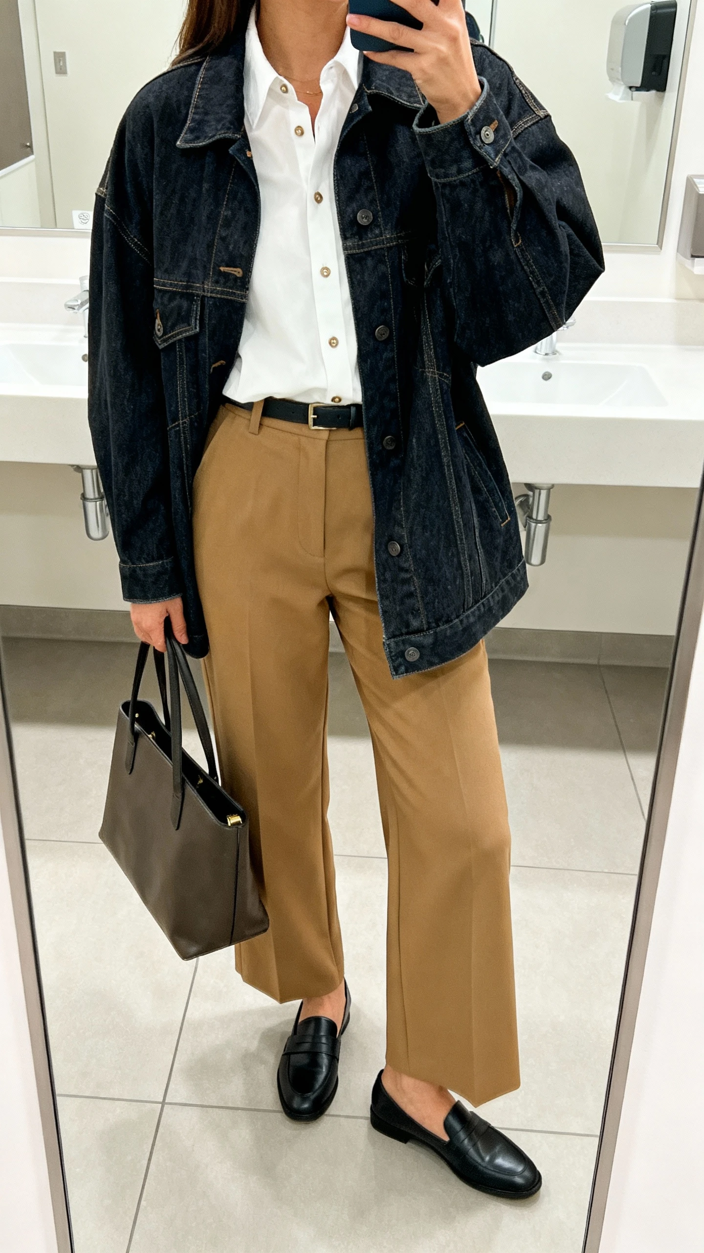 iPhone office-bathroom mirror selfie of a woman wearing an oversized dark-wash denim jacket over a crisp white button-down tucked into high-waisted wide-leg camel trousers, black loafers, and a structured tote; face not visible, bright indoor lighting, natural iPhone photo quality.