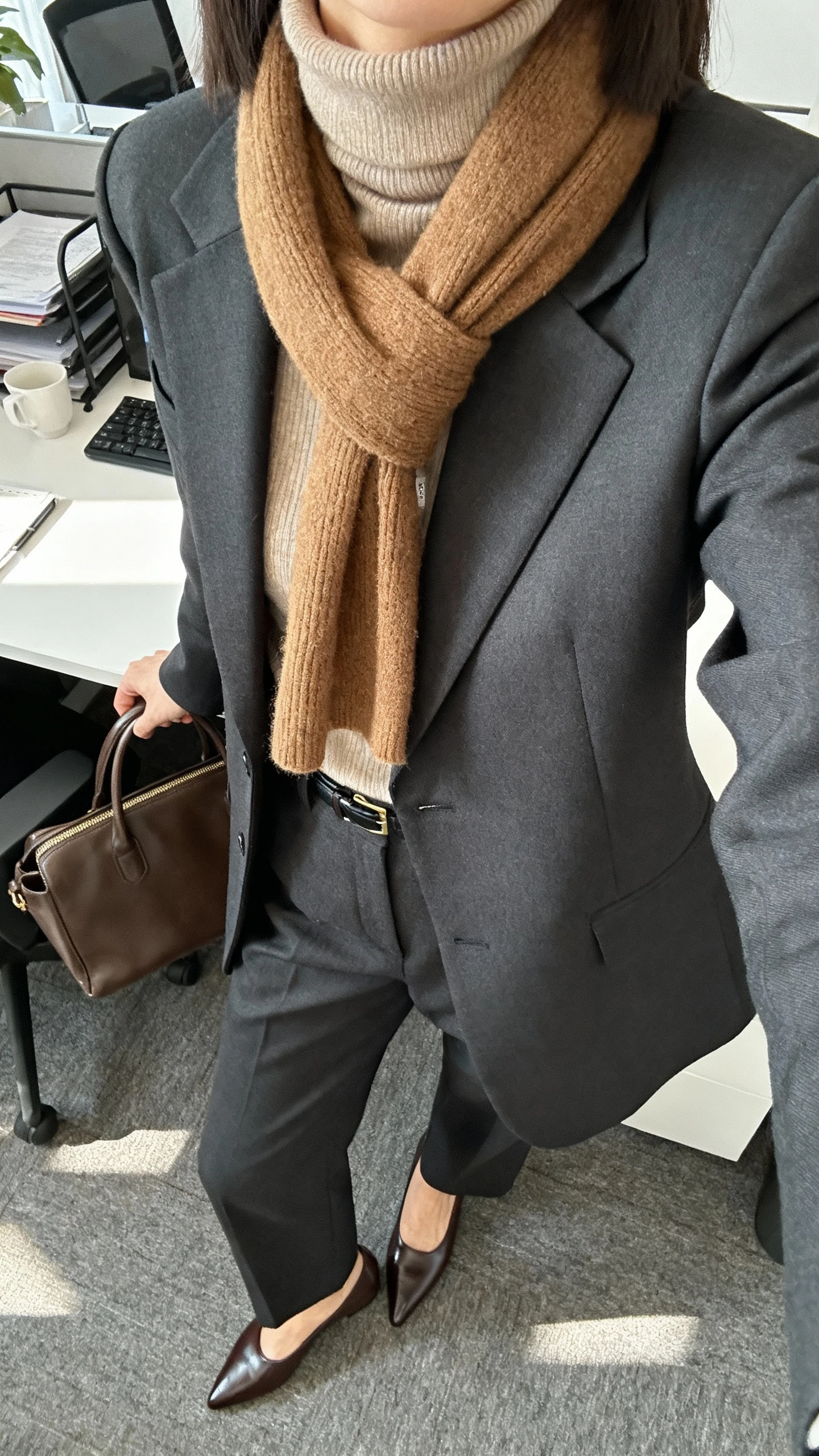 iPhone office desk selfie cropped at neck showing a woman in a tailored charcoal suit (blazer + trousers) layered with a camel cashmere turtleneck and scarf, pointed loafers, leather brief tote, face not visible, morning office light, natural iPhone photo quality.