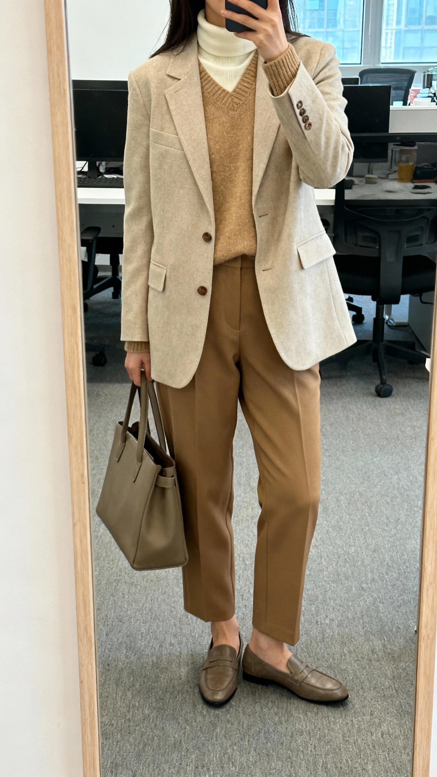 iPhone office mirror selfie of a woman wearing a beige blazer layered over a camel knit, cream turtleneck peeking at the collar, tan tailored trousers, loafers, structured tote, face not visible, cropped at shoulders, morning office light, natural iPhone photo.