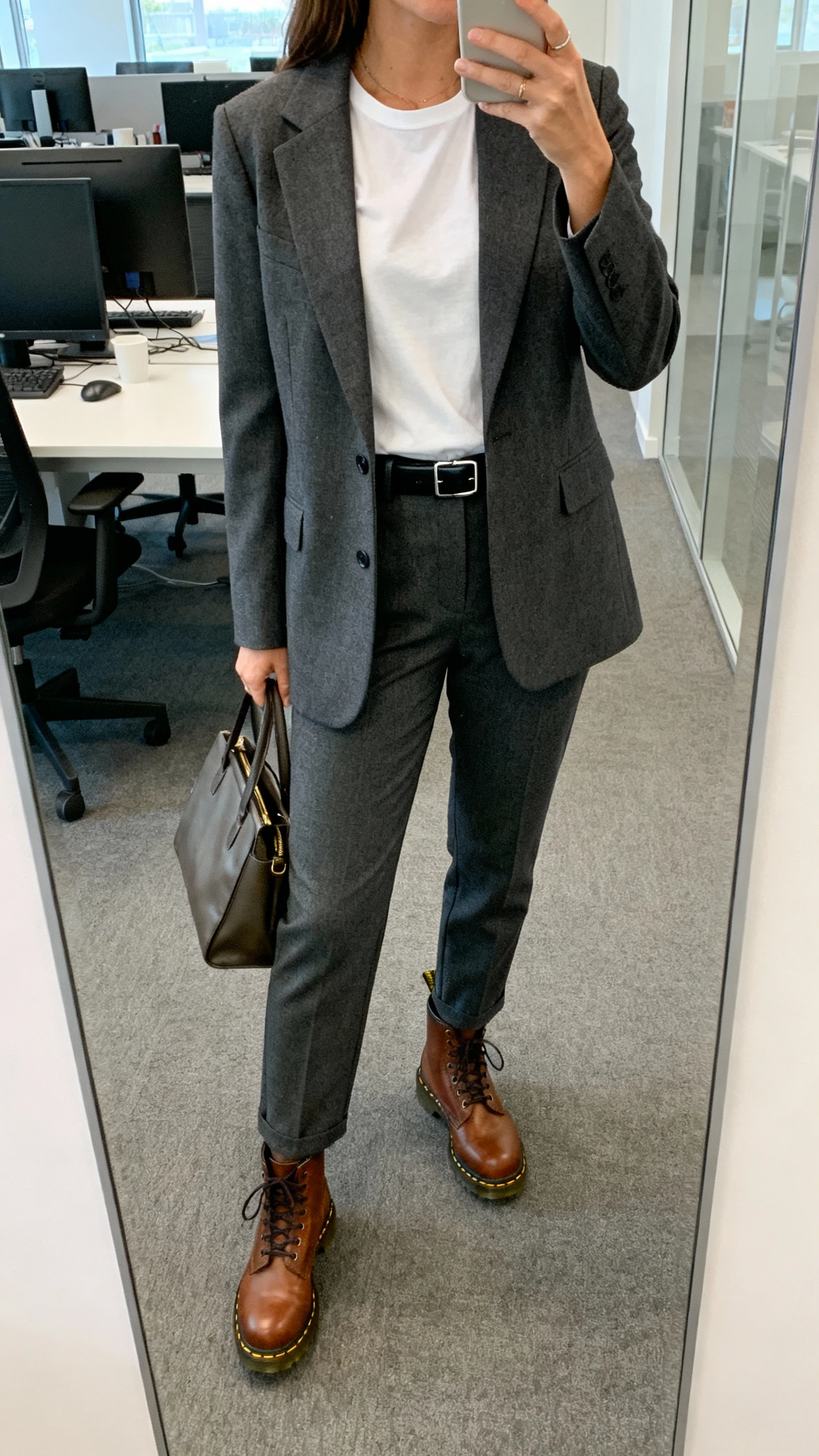 iPhone office mirror selfie of a woman wearing a tailored charcoal blazer and matching trousers, crisp white tee, brown Doc Martens, slim black belt, and structured tote; face not visible, cropped at shoulders, morning office light, natural iPhone photo.