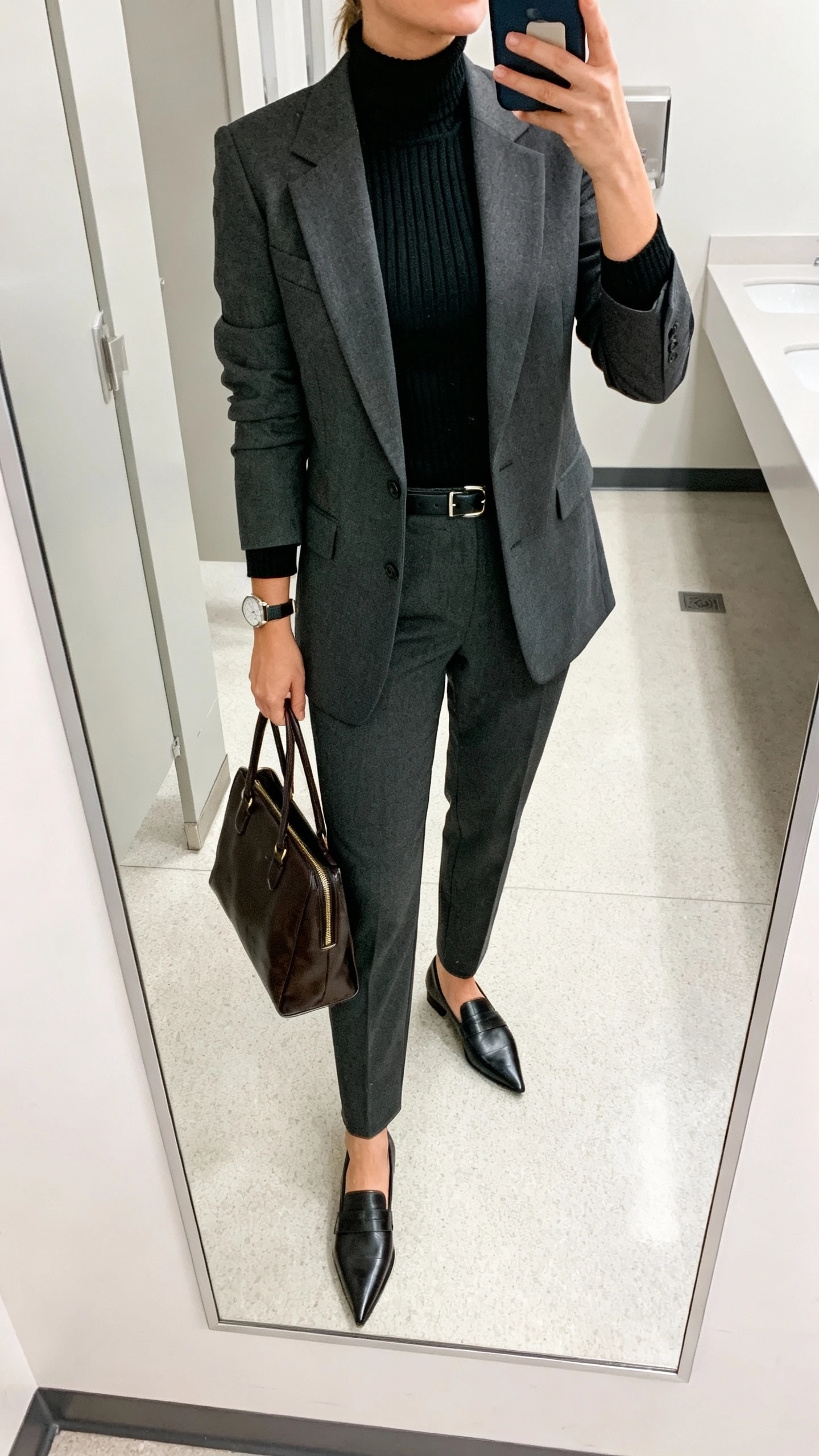 iPhone office-mirror selfie of a woman wearing tailored suiting with a turtleneck: sharp charcoal blazer and matching trousers, black ribbed turtleneck, pointed-toe loafers, leather work tote, slim belt, watch, face not visible, cropped at shoulders, office restroom mirror, bright indoor lighting, natural iPhone quality.