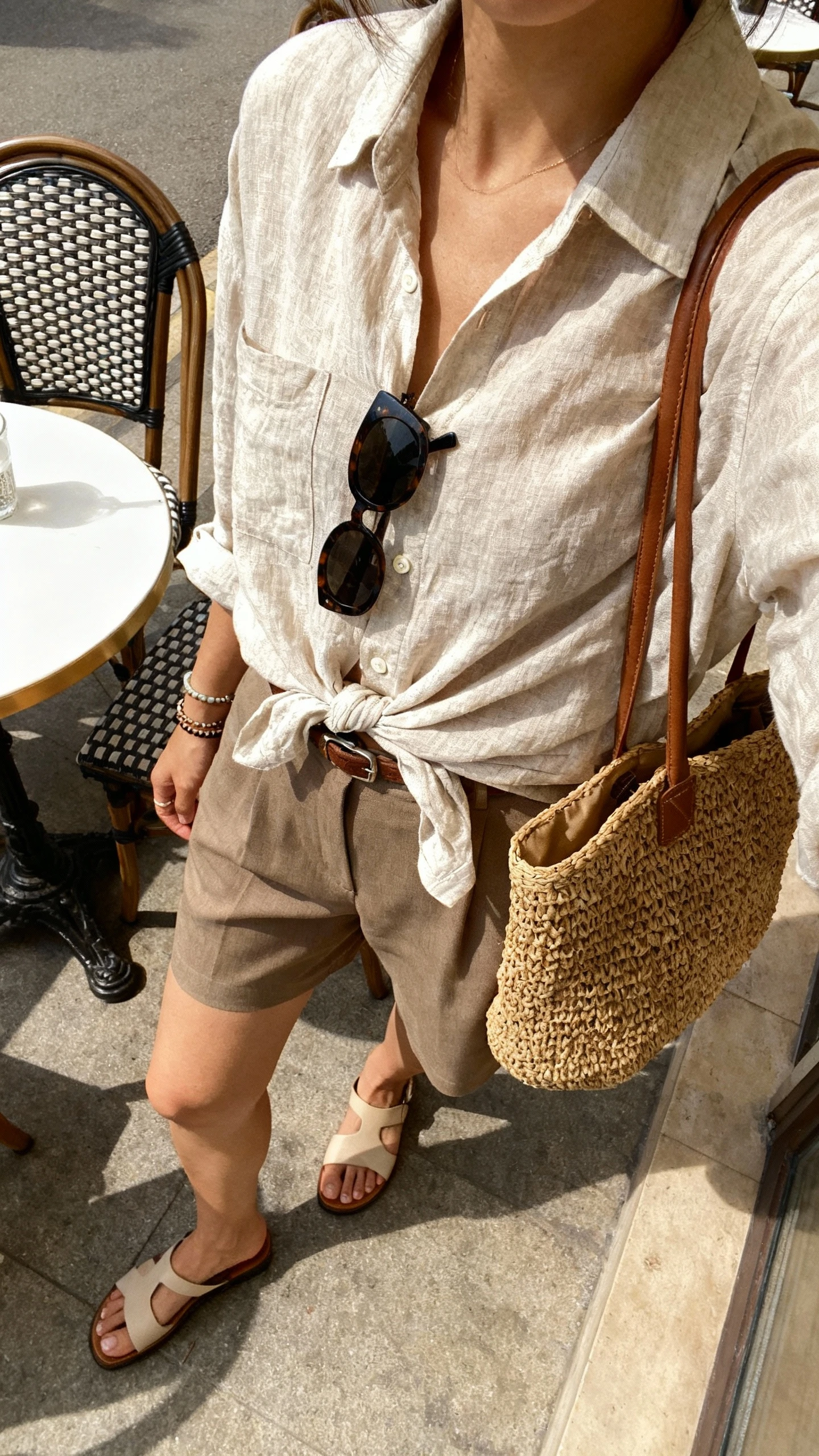 iPhone patio table selfie (downward angle) of a woman wearing a relaxed linen button-down half-tucked into tailored shorts, flat sandals, woven tote, sunglasses, and simple bracelets, face not visible, sunny cafe patio with natural light, natural iPhone photo quality.