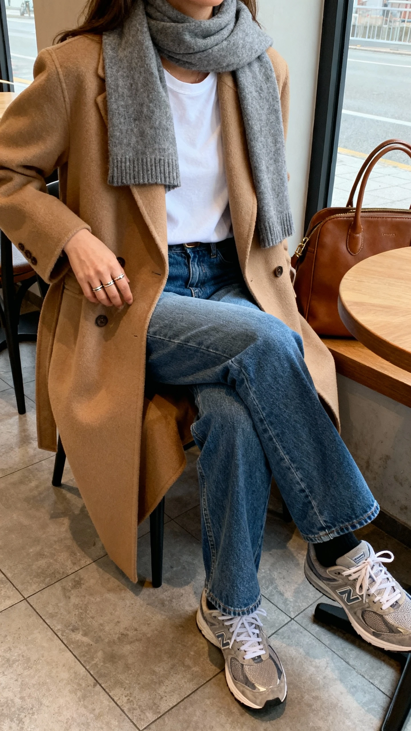 iPhone seated cafe selfie of a woman in casual-cool denim with elevated layers: straight blue jeans, crisp white tee, tailored camel coat, gray cashmere scarf, sneakers, leather tote, subtle rings, face not visible, cropped at shoulders while seated by window, natural daylight, casual iPhone photo.