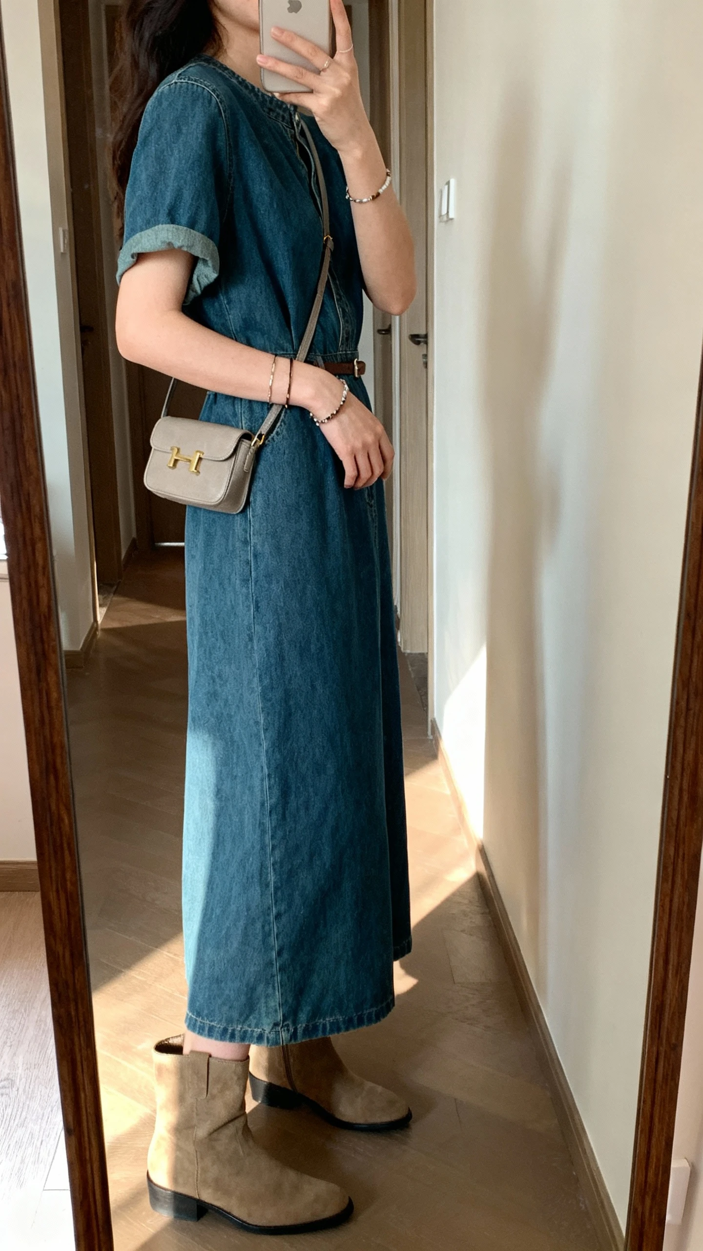 iPhone selfie from a side angle showing a woman in an effortless denim dress with mid-calf boots, small shoulder bag, subtle bracelets, face not visible, hallway mirror in an apartment, soft afternoon light, casual iPhone aesthetic.