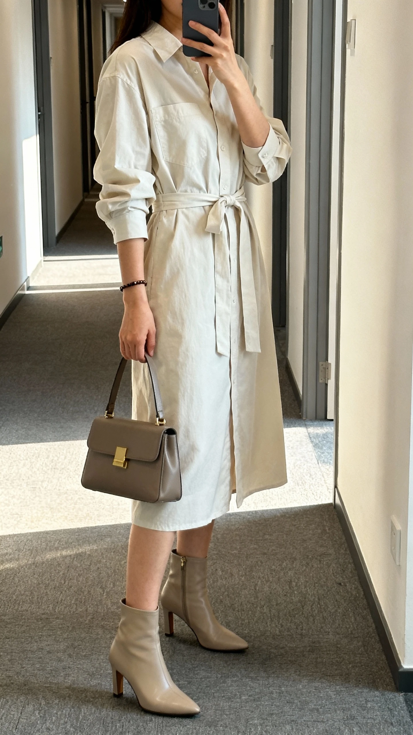 iPhone side-angle hallway selfie of a woman wearing an elevated shirt dress with a waist tie, heeled ankle boots, structured satchel, simple bracelet, face not visible, morning office corridor light, natural iPhone photo quality.