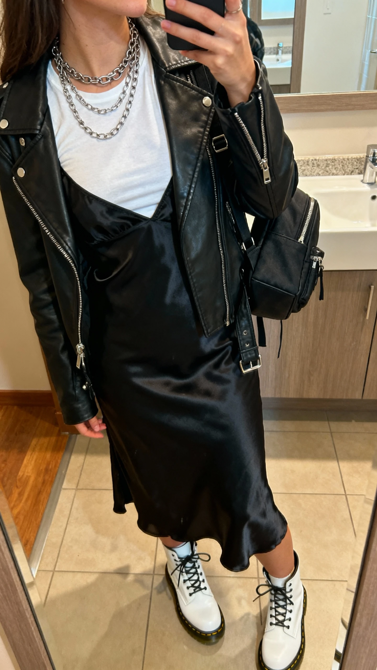 iPhone side-angle mirror snap of a woman in a grunge slip dress: black satin slip layered over a fitted white tee, black leather moto jacket, white Doc Martens, layered chains, black mini backpack; face not visible, bathroom mirror, warm indoor light, casual iPhone aesthetic.