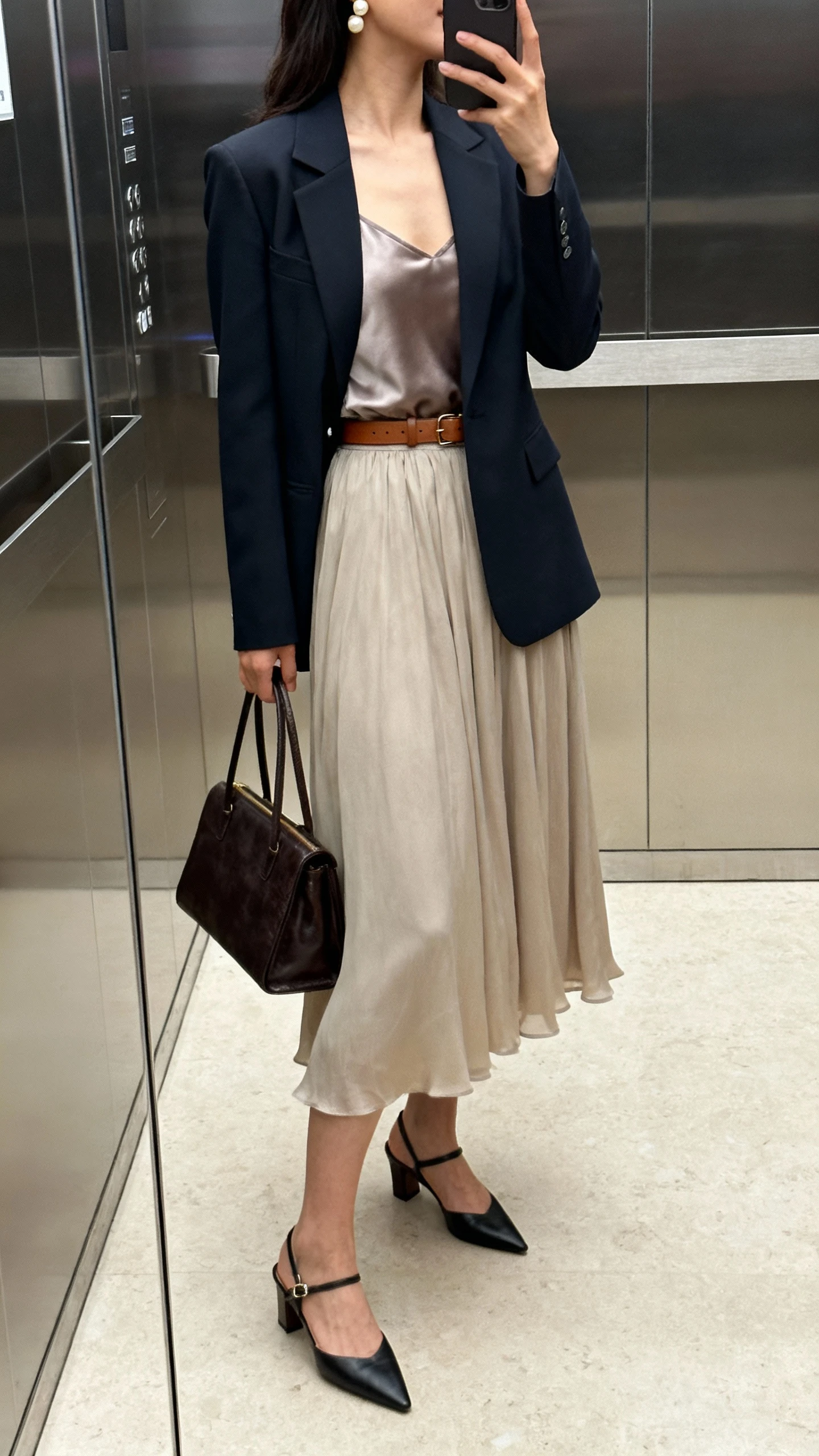 iPhone side-angle selfie in an office elevator mirror of a woman wearing a blazer + midi skirt combo: fitted blazer, flowy midi skirt, silky camisole, slingback heels, pearl studs, slim leather belt, structured tote; face not visible, indoor lighting, natural iPhone photo quality.