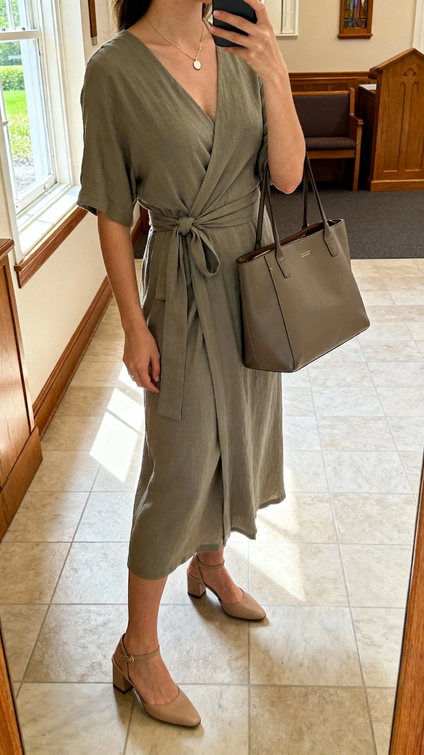 iPhone side-angle selfie of a woman in a modest wrap dress with slingback heels, dainty pendant necklace, structured tote, face not visible, taken in a church foyer by a window, natural morning light, casual iPhone photo.