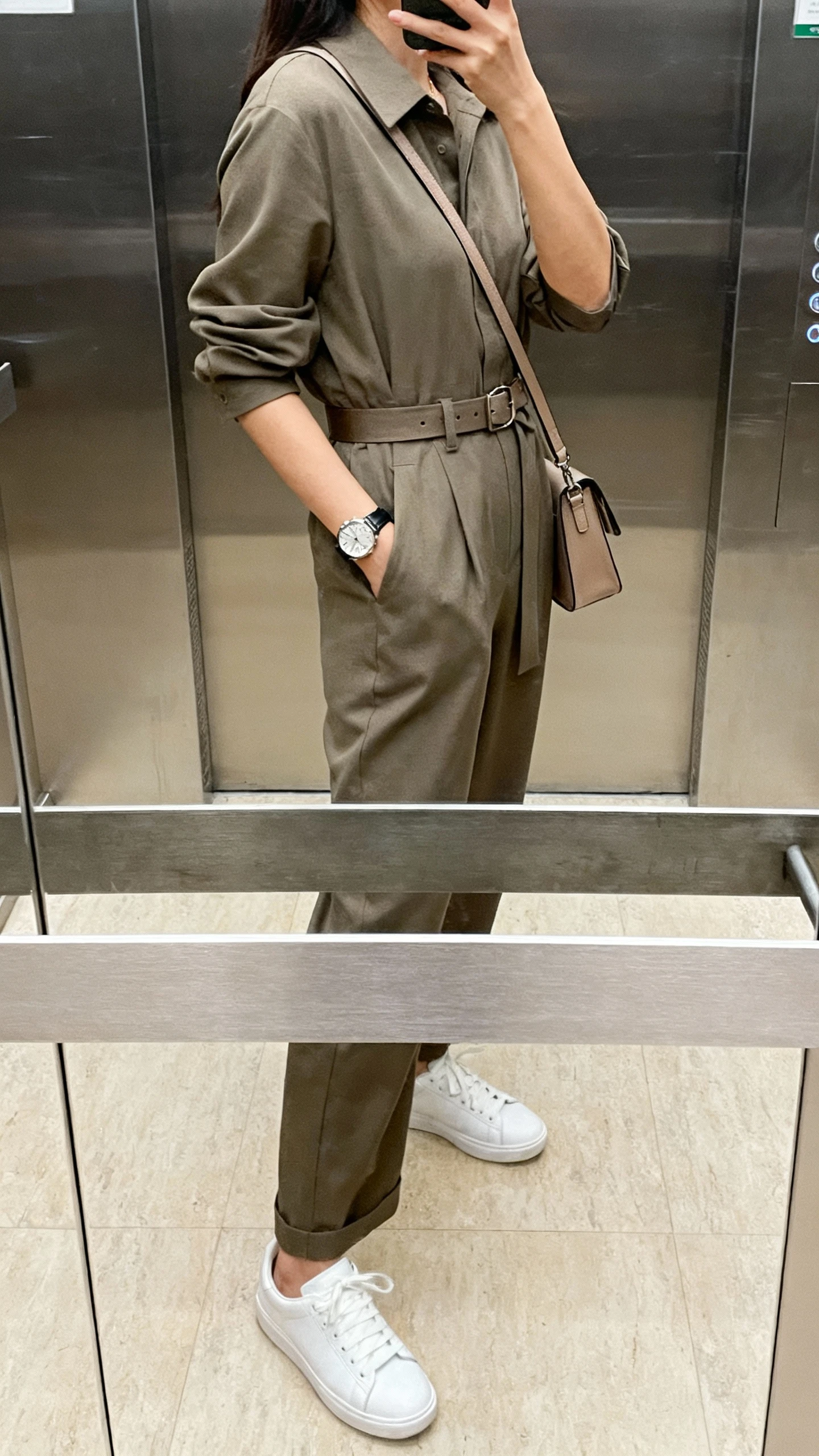 iPhone side-angle selfie of a woman in a smart-casual jumpsuit cinched with a belt, white sneakers, watch, crossbody bag, face not visible, elevator mirror, indoor lighting, natural iPhone aesthetic.