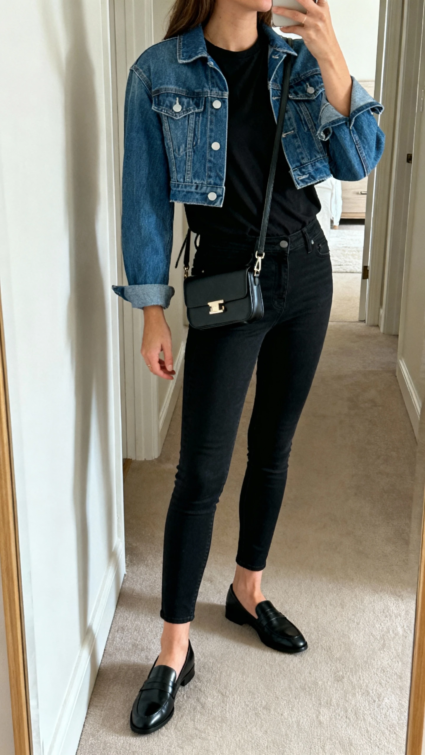 iPhone side-angle selfie of a woman in an all-black base (black tee and skinny jeans) with a cropped blue denim jacket, black loafers, mini shoulder bag, face not visible, hallway mirror, indoor lighting, natural iPhone aesthetic