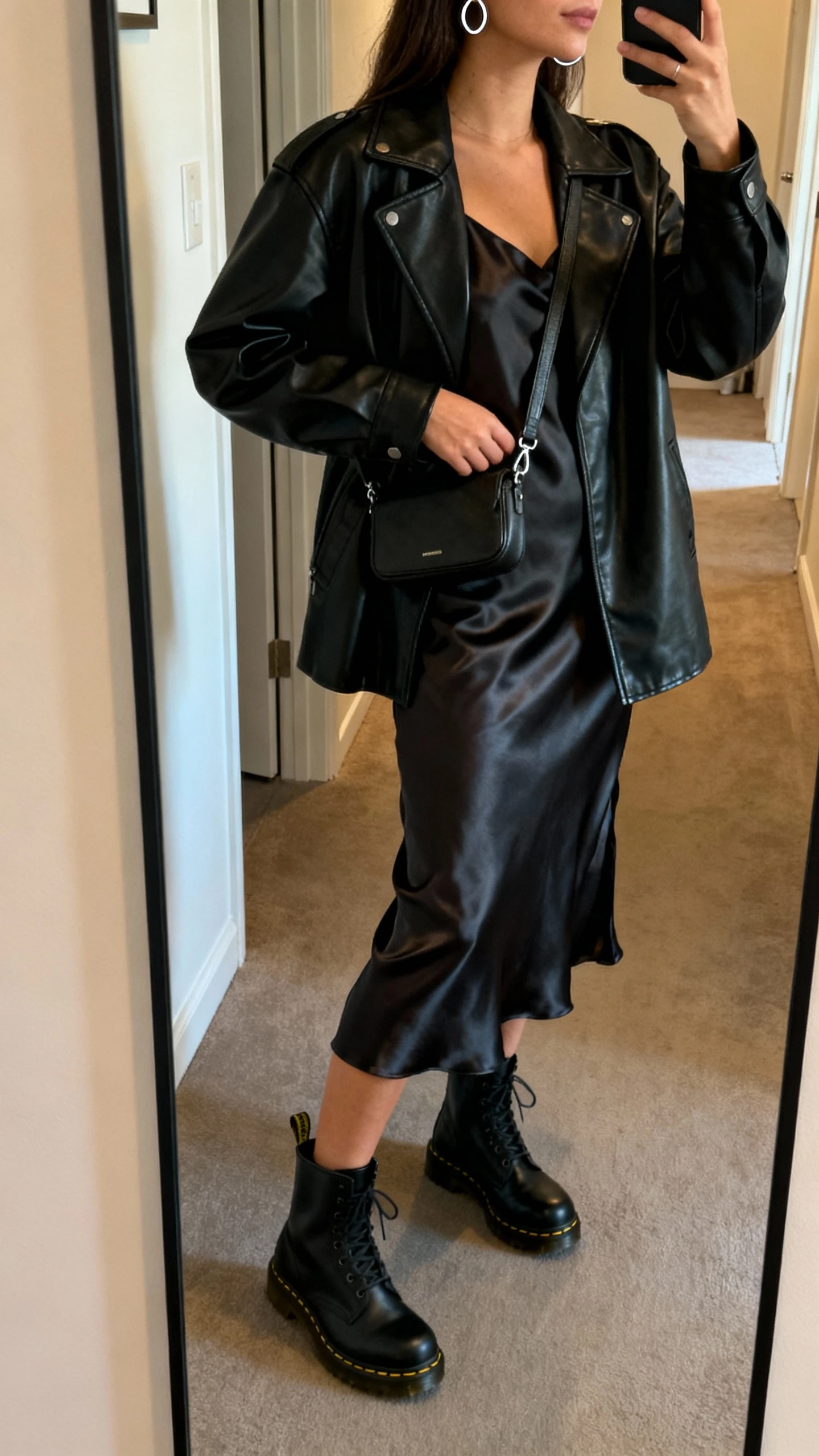 iPhone side-angle selfie of a woman wearing a black leather jacket over a satin slip dress, black Doc Martens, minimal silver hoops, small crossbody bag, face not visible, hallway mirror in an apartment, warm indoor lighting, natural iPhone photo quality.