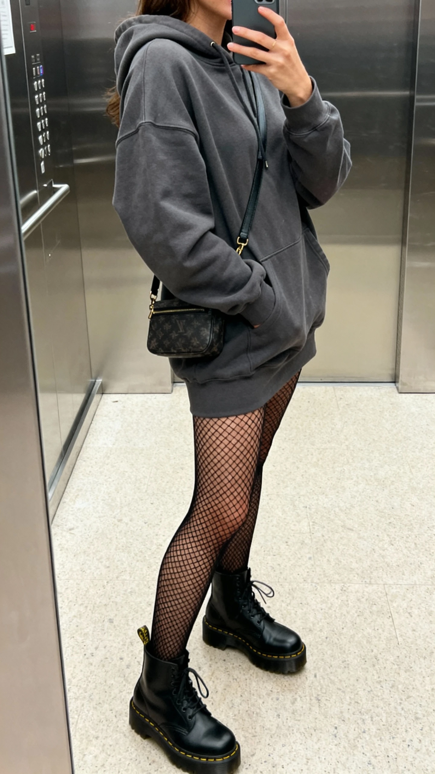 iPhone side-angle selfie of a woman wearing a charcoal hoodie dress, black fishnet tights, black platform Doc Martens, crossbody bag, face not visible, elevator mirror after shopping, afternoon indoor lighting, natural iPhone photo quality.