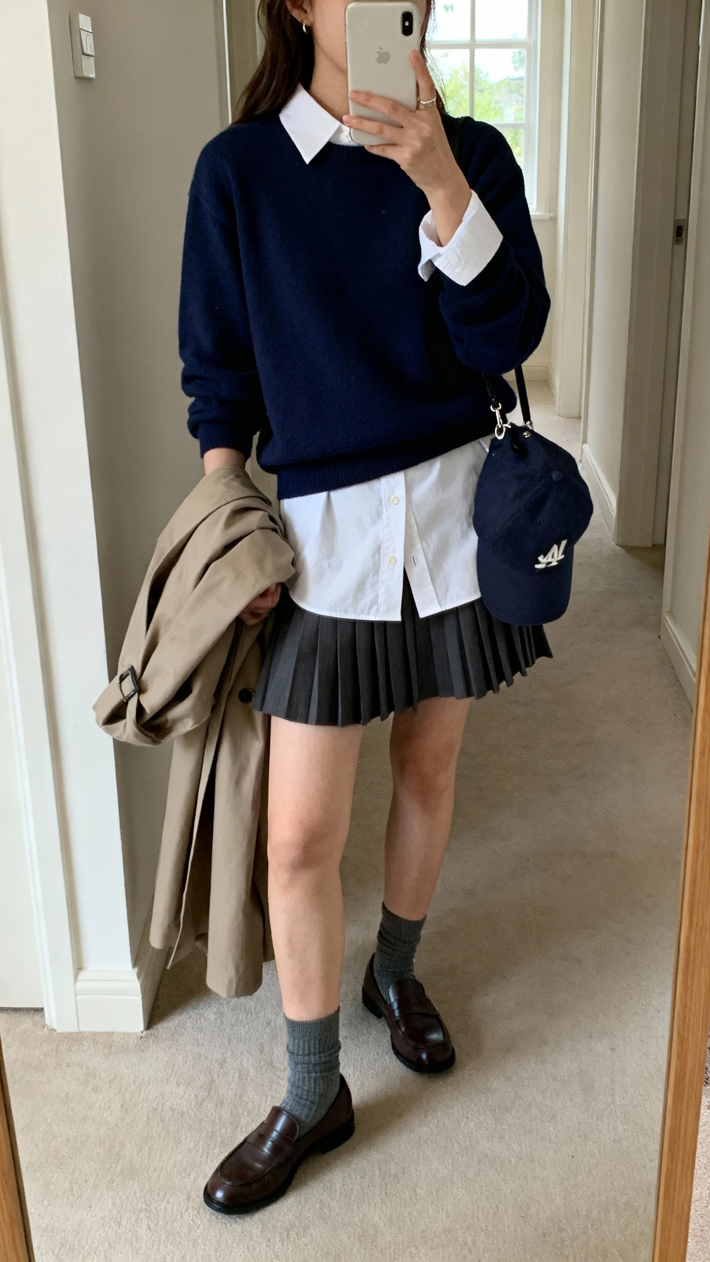 iPhone side-angle selfie of a woman wearing a crisp white button-down layered under a navy crewneck sweater, pleated mini skirt, loafers with socks, slouchy trench over arm, baseball cap clipped to bag for street vibe, face not visible, hallway mirror, natural window light, iPhone photo quality.