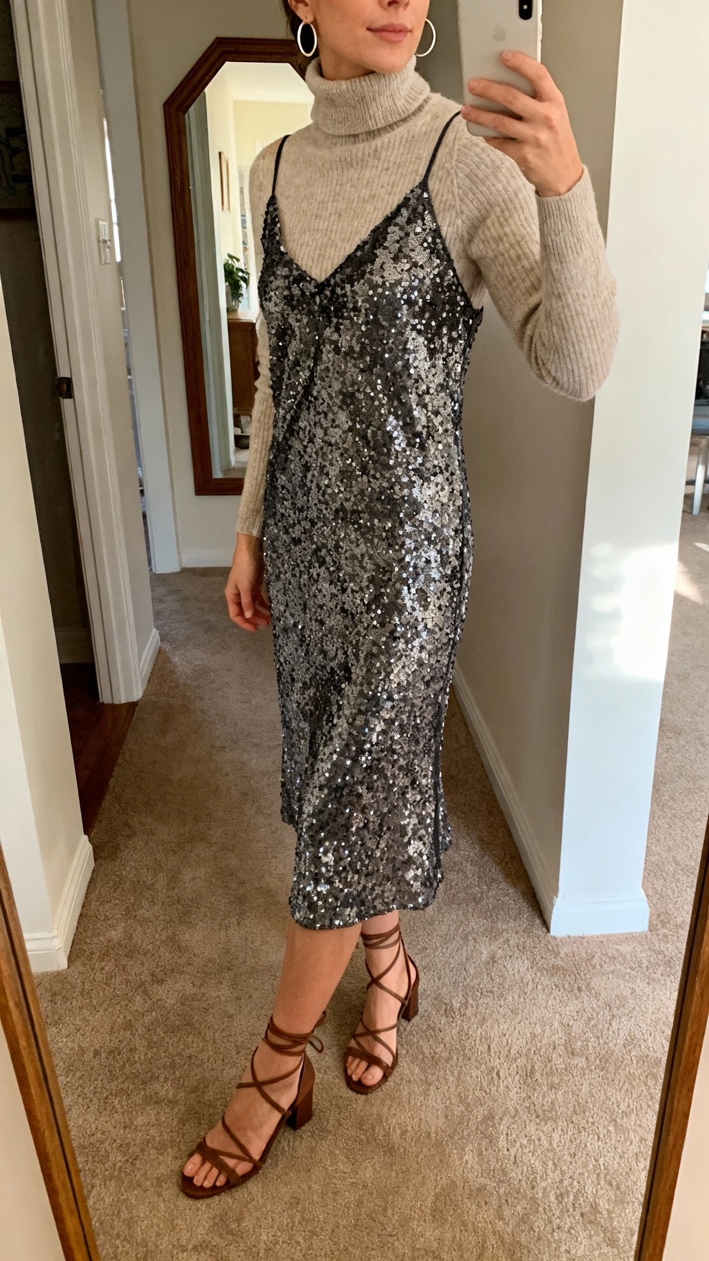 iPhone side-angle selfie of a woman wearing a sequin slip layered over a cashmere turtleneck, minimal hoops and strappy heels, face not visible, hallway mirror at home, warm indoor lighting, natural iPhone photo quality.