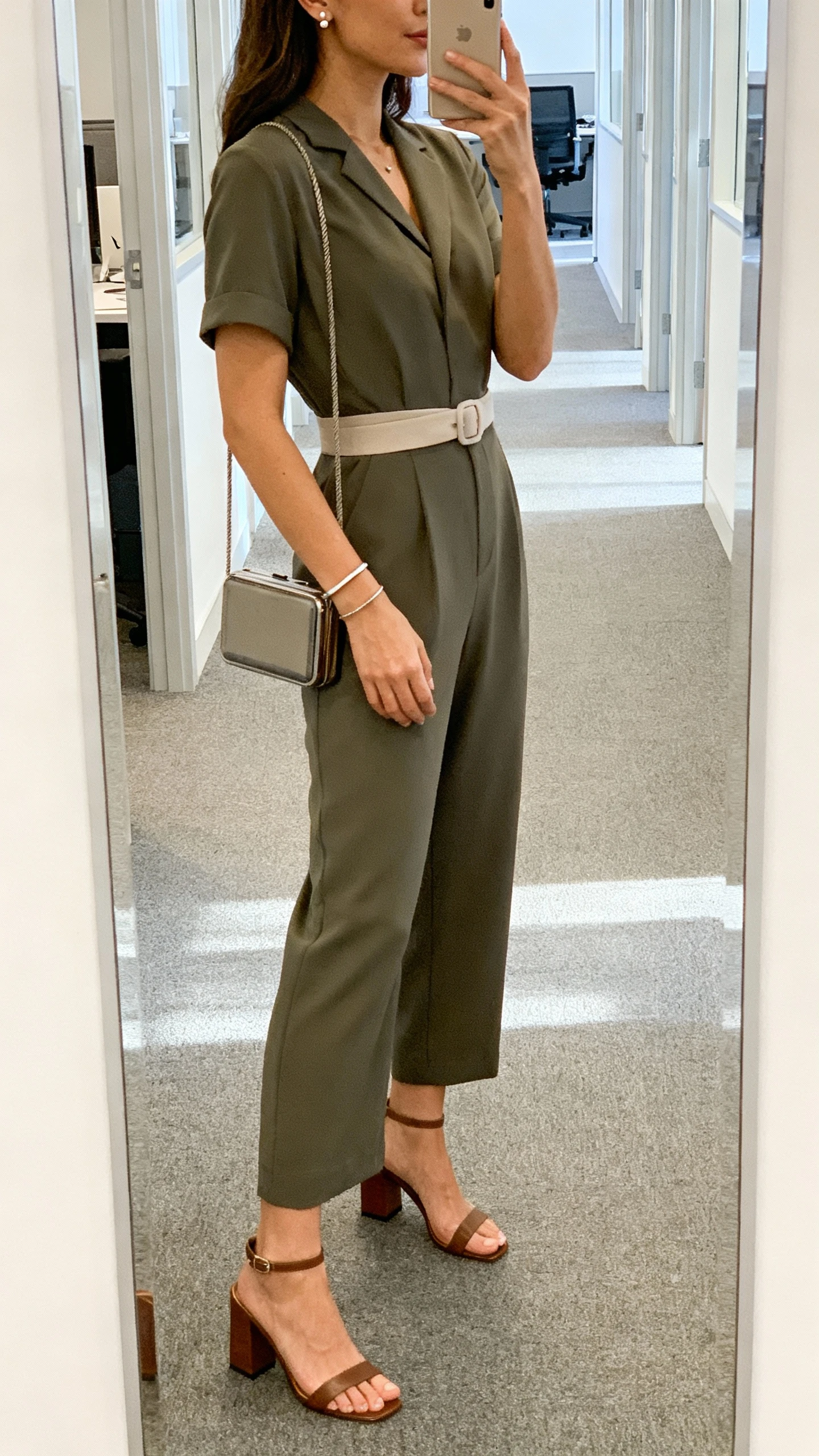 iPhone side-angle selfie of a woman wearing a tailored short-sleeve jumpsuit with a soft waist belt, block-heel sandals, minimal stud earrings and thin cuff bracelet, compact crossbody; office hallway mirror, bright natural light, face not visible, casual iPhone look.