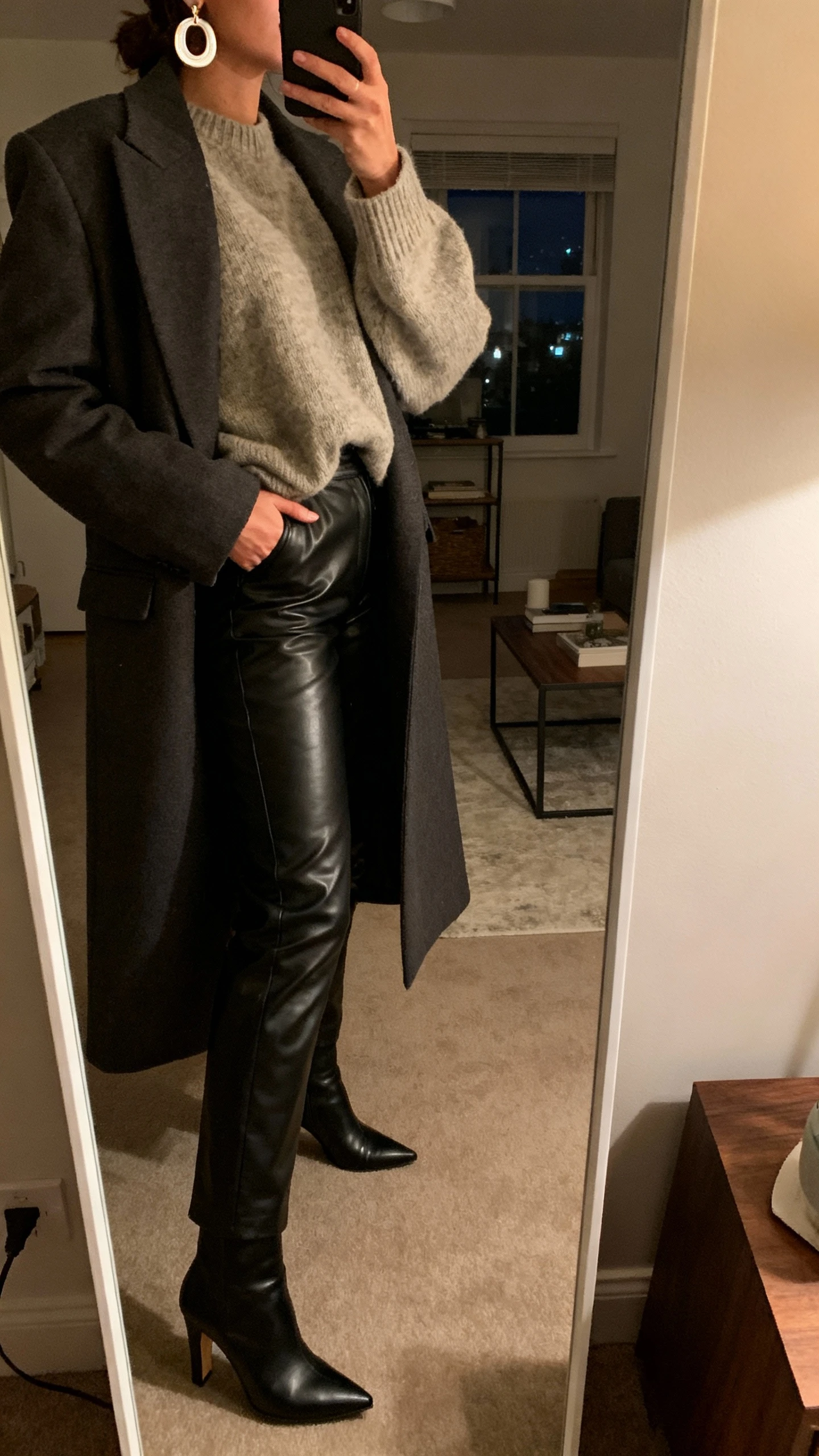 iPhone side mirror selfie of a woman in faux leather trousers, slouchy cashmere sweater, sleek heeled ankle boots, long tailored coat, bold earrings; apartment full-length mirror, face not visible, evening indoor lighting, natural iPhone aesthetic.