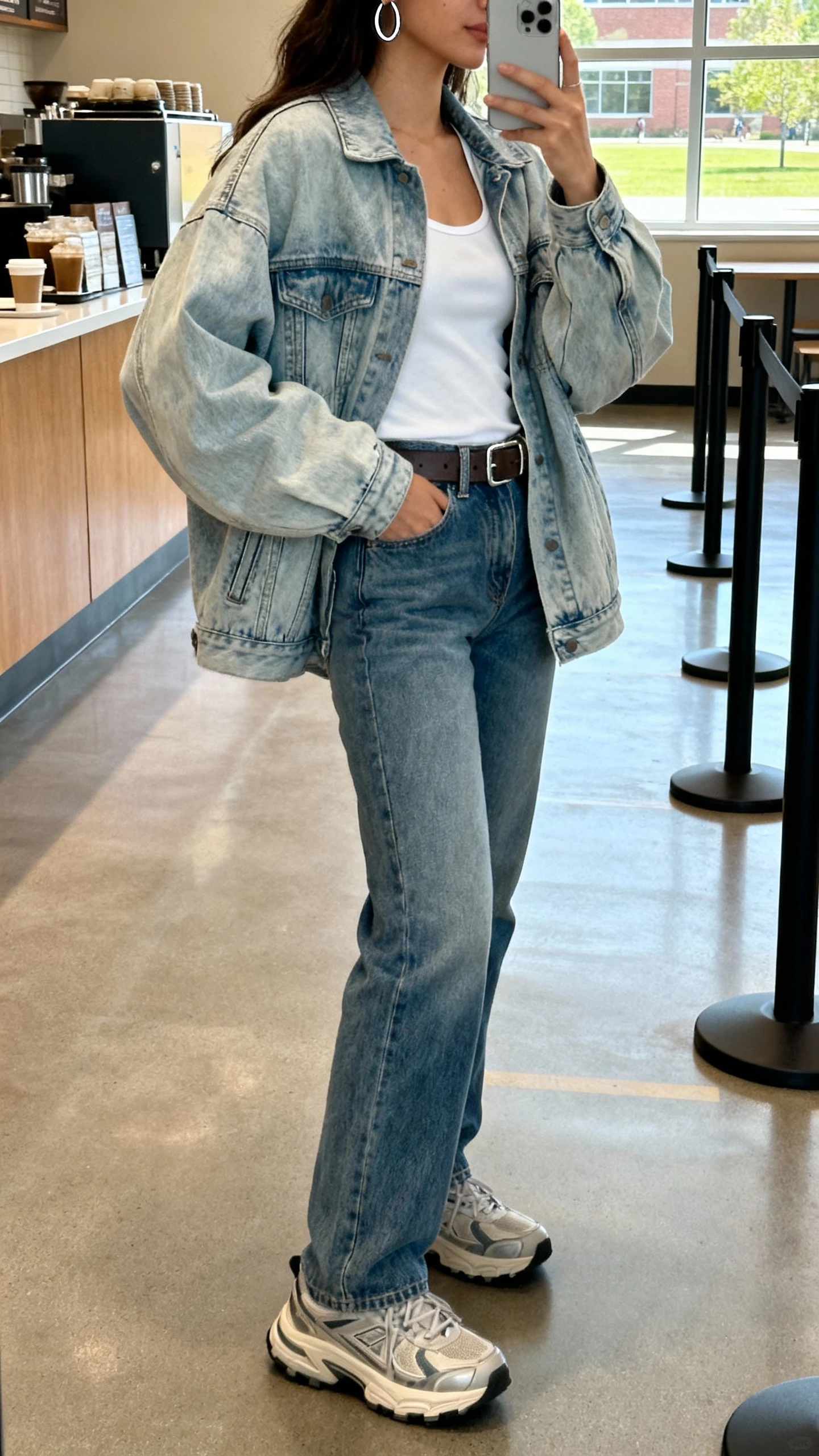 iPhone side-view selfie of a woman in denim-on-denim: light-wash oversized denim jacket with a white tank and mid-wash straight jeans, belt, chunky sneakers, silver hoops, face not visible, campus café line, daylight from window, casual iPhone aesthetic.