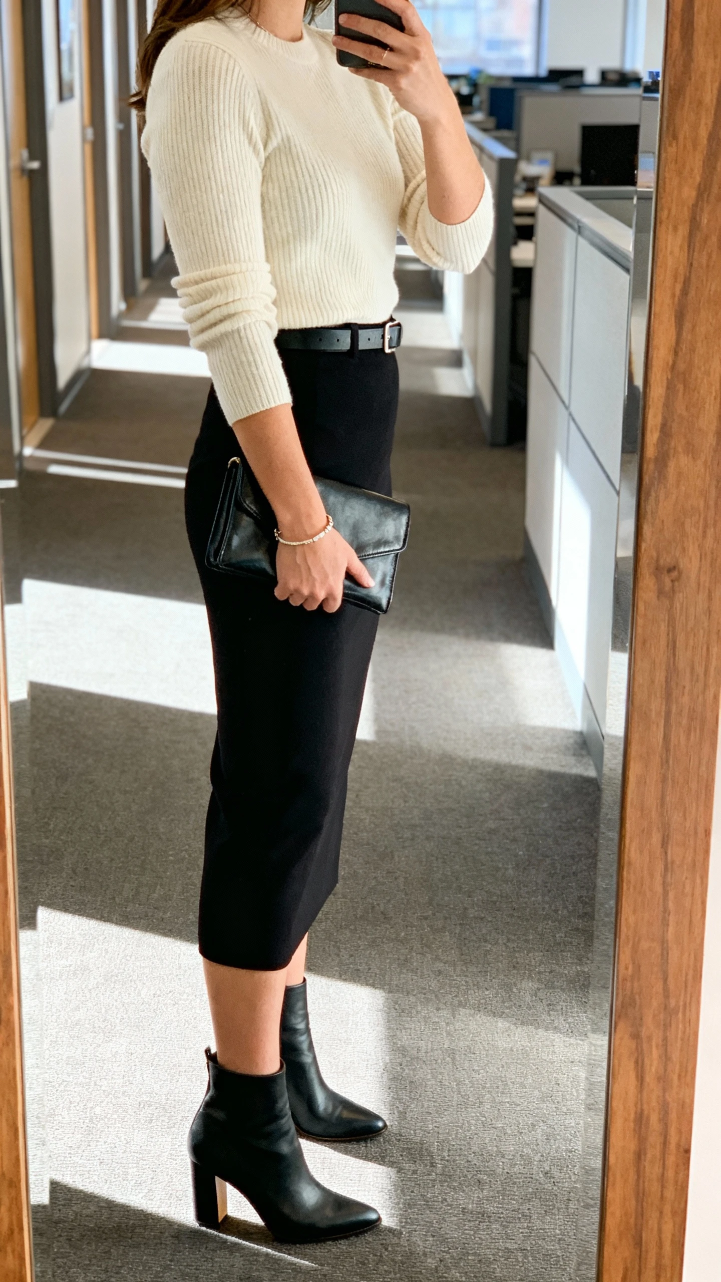 iPhone side-view selfie of a woman wearing a column skirt and knit combo: black column skirt with a fitted fine-knit sweater in cream, heeled ankle boots, minimalist belt, delicate bracelet, compact leather clutch, face not visible, corridor mirror at work, natural afternoon light, iPhone photo quality.
