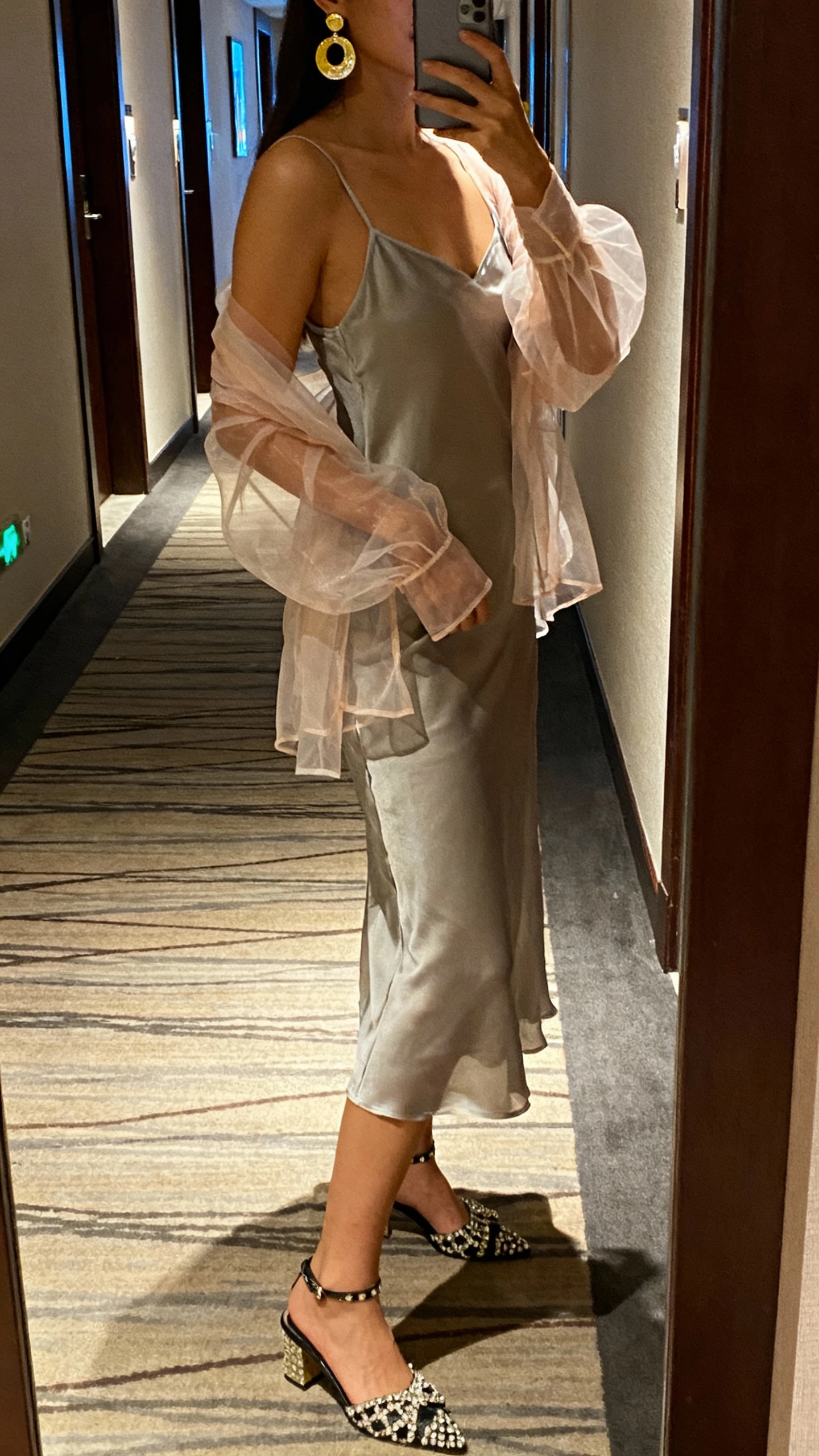 iPhone side-view selfie of a woman wearing a slip dress under a sheer organza top with statement heels and bold earrings, face not visible, hotel hallway mirror, evening indoor lighting, natural iPhone photo quality.
