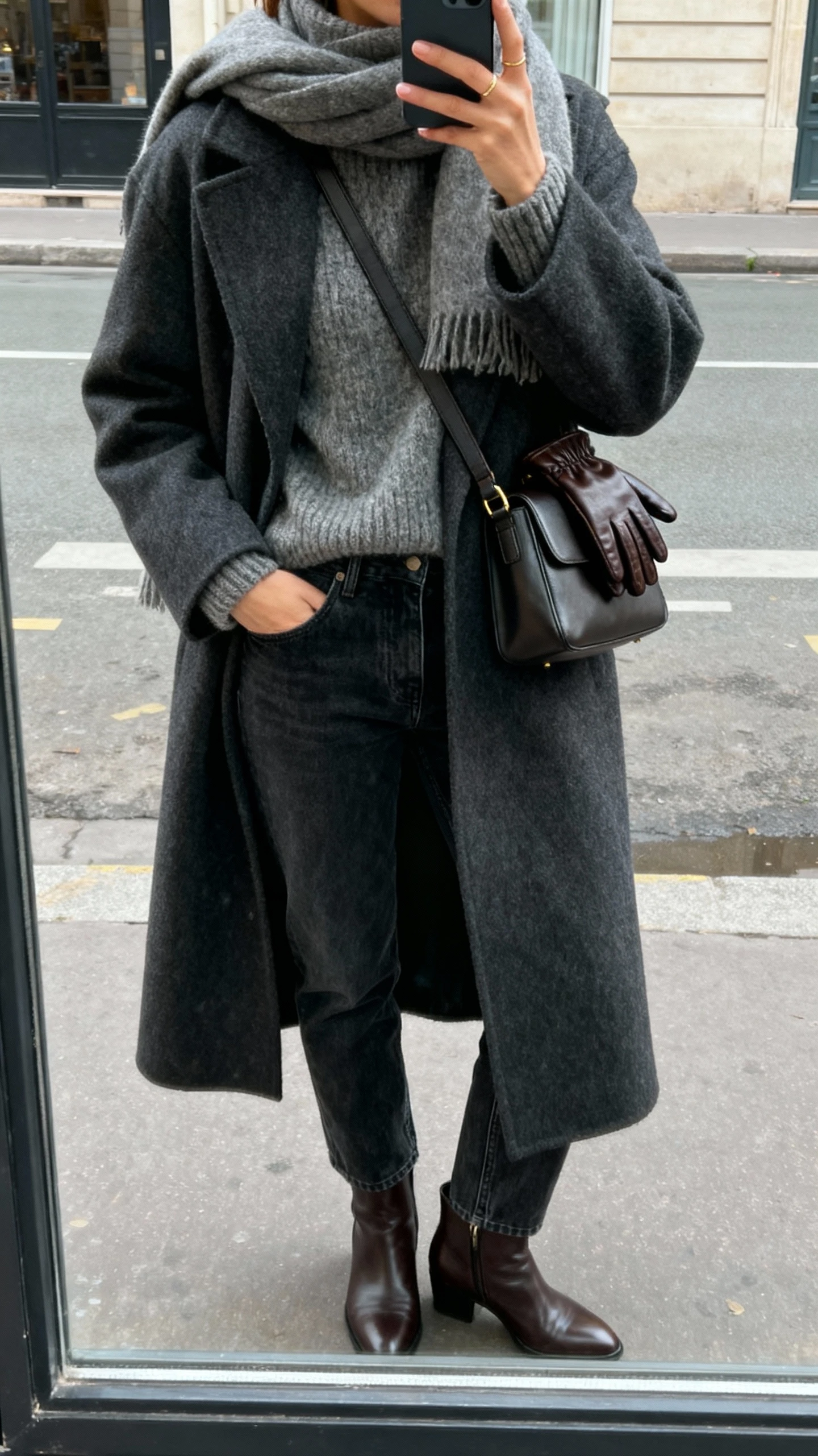 iPhone street mirror-window selfie of a woman in a long wool coat (charcoal), chunky crewneck sweater, straight-leg dark-wash jeans, leather ankle boots, cashmere scarf, leather gloves tucked into a crossbody/structured shoulder bag; face not visible, overcast natural daylight, casual iPhone photo.