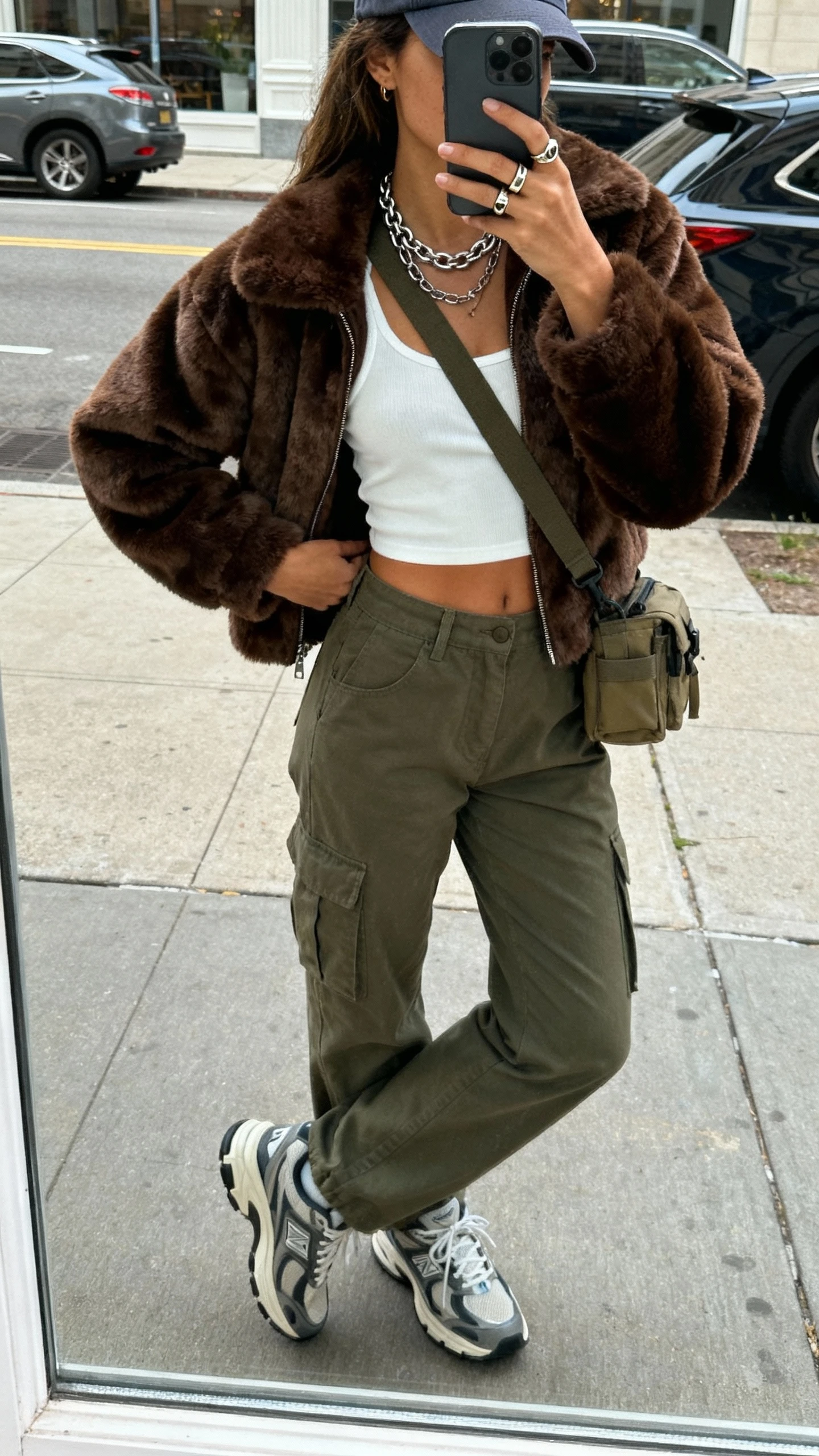 iPhone street mirror window selfie of a woman in a chocolate faux fur bomber, cream cropped tank, high-waisted olive cargo pants, chunky retro sneakers, baseball cap, crossbody utility bag, layered chain necklaces and a signet ring; face not visible, side angle, daylight, natural iPhone photo quality.