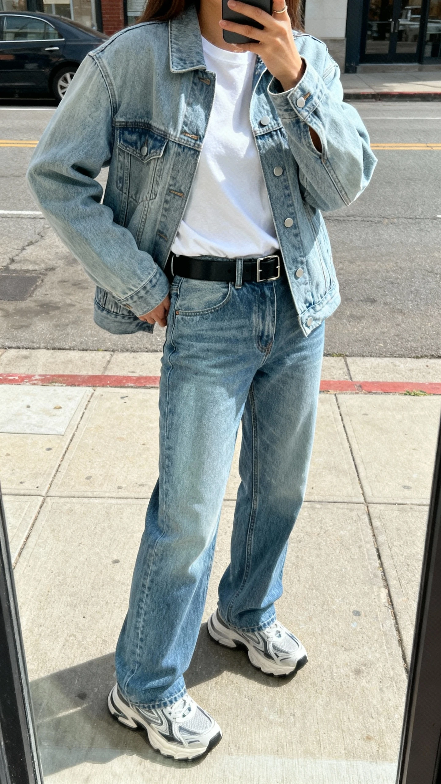 iPhone street-mirror window selfie of a woman in denim on denim: light-wash jacket over mid-wash straight jeans, white tee, black belt, chunky sneakers, face not visible, urban sidewalk reflection, natural daylight, iPhone photo quality