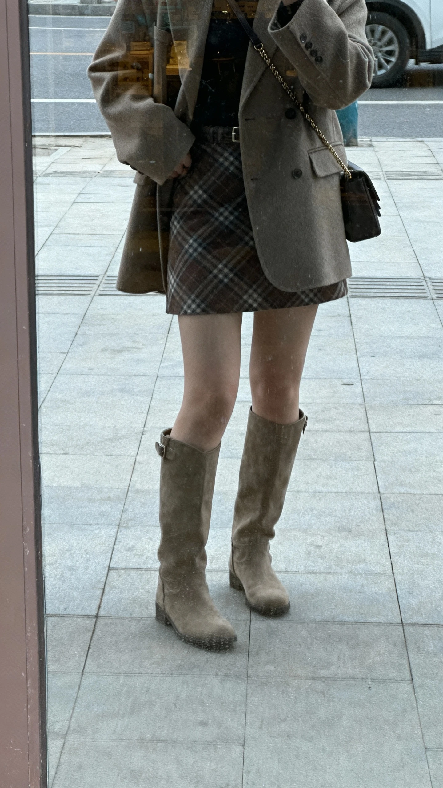 iPhone street-side mirror reflection (storefront glass) of a woman wearing a plaid mini skirt, tall boots, and a polished coat, crossbody bag, face not visible, overcast daylight, natural iPhone photo quality.
