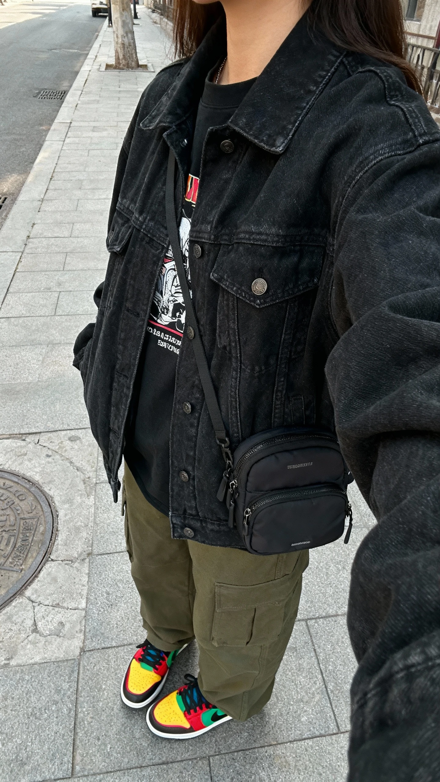 iPhone street-side selfie from a side angle showing a woman wearing an oversized black denim jacket, olive cargo pants, bold multicolor sneakers, graphic tee, and a mini backpack; natural daylight, city sidewalk, face not visible, natural iPhone photo quality.