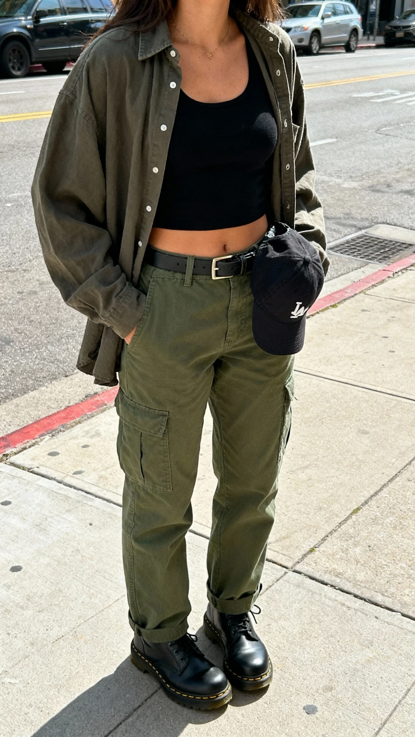iPhone street-style selfie from chest down of a woman wearing olive cargo pants, black cropped tank, slouchy unbuttoned overshirt, black Doc Martens, baseball cap clipped to belt loop, face not visible, city sidewalk, bright daylight, natural iPhone photo quality.
