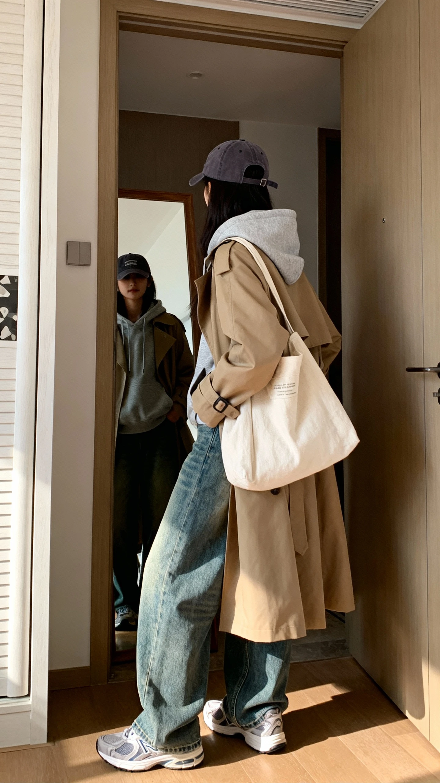 iPhone back/side view of a woman wearing baggy jeans, a hoodie layered under a classic trench coat, comfy sneakers, canvas tote, baseball cap, face not visible, entryway mirror, natural daylight, iPhone photo quality.
