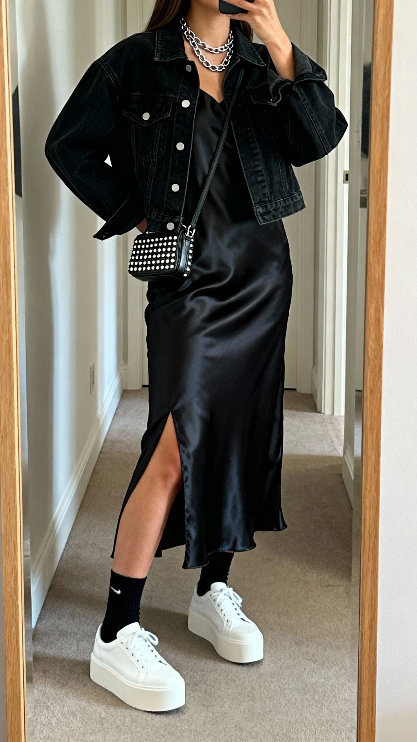 iPhone back-view mirror selfie of a woman wearing a black denim jacket over a black satin slip dress with a slit, black crew socks, white platform sneakers, layered silver chain necklace, and a small studded mini bag, face not visible, hallway full-length mirror, natural daylight, iPhone photo quality.