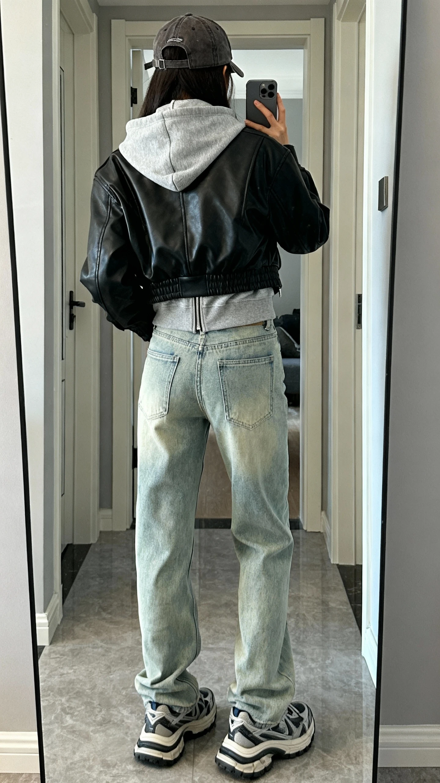 iPhone back-view mirror selfie of a woman wearing light-wash mom jeans, a gray zip hoodie layered under a black leather jacket, chunky sneakers, and a baseball cap, face not visible, entryway mirror, natural daylight, iPhone photo quality.