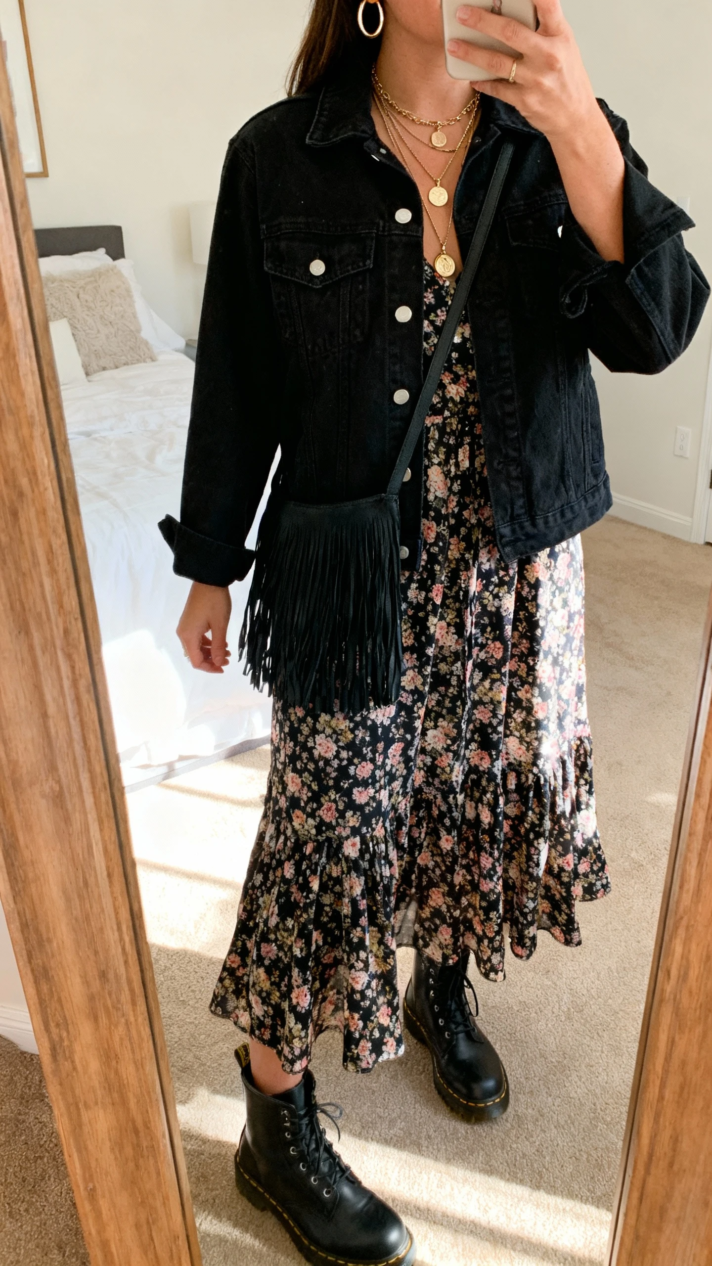iPhone back-view mirror selfie of a woman wearing a black denim jacket over a flowy floral midi dress, black combat boots, layered pendant necklaces, hoop earrings, and a black fringe crossbody, face not visible, bedroom mirror, warm afternoon light, iPhone photo quality.