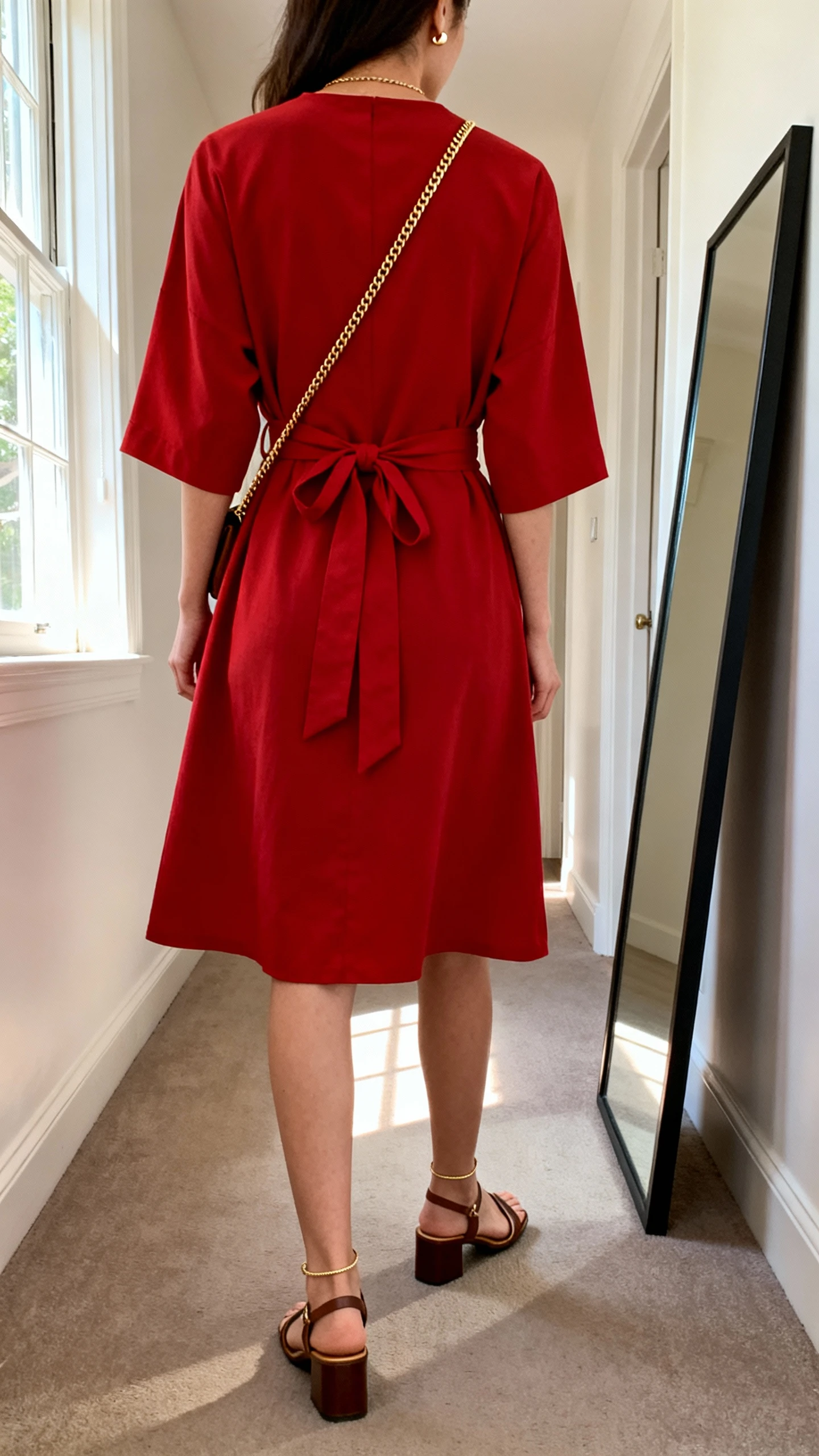 iPhone back view of a woman wearing a red wrap dress with tie waist, low block-heel sandals, thin gold chain and small stud earrings, crossbody bag, face not visible, hallway full-length mirror, morning window light, iPhone photo quality