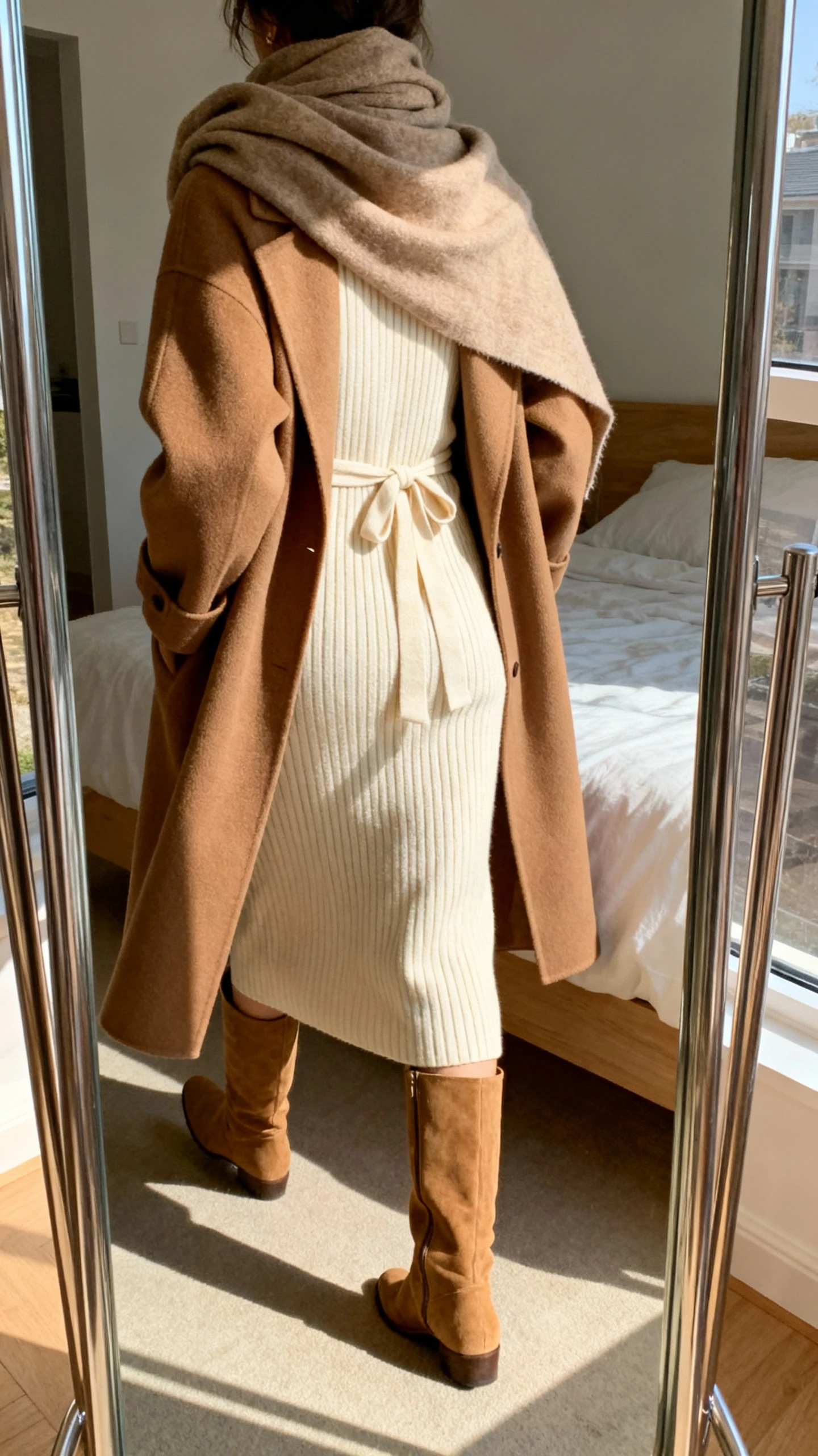 iPhone back view of a woman wearing a soft brown coat over a ribbed cream knit midi dress with a tie waist, tan knee-high boots, and a slouchy scarf, face not visible, walking pose in bedroom mirror, warm afternoon light, iPhone photo quality.