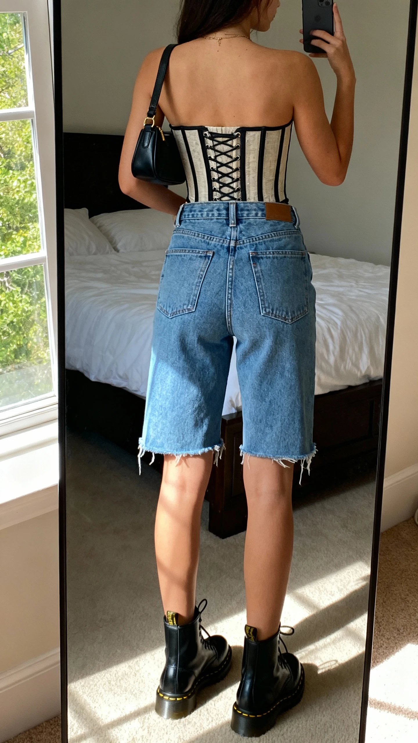 iPhone back view of a woman wearing high-rise denim cutoffs, a structured corset top, black Doc Martens, and a small shoulder bag, face not visible, bedroom mirror, afternoon window light, iPhone photo quality.