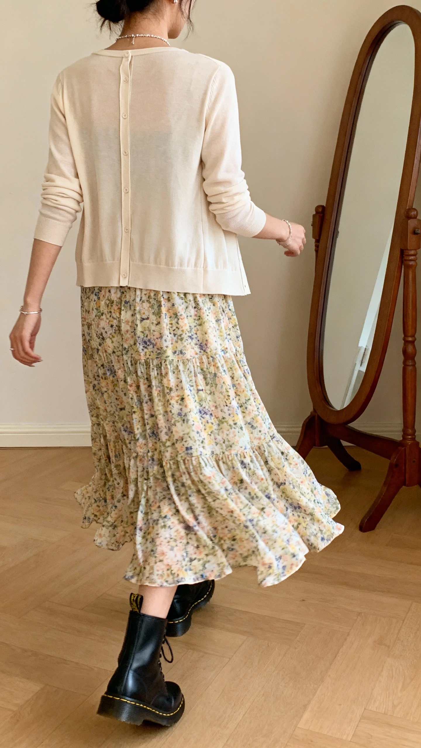 iPhone back view photo of a woman wearing a floaty floral skirt styled like a dress with a slim cardigan layered on top, black Doc Martens and delicate jewelry, face not visible, walking pose by a full-length mirror, warm indoor lighting, iPhone photo quality.