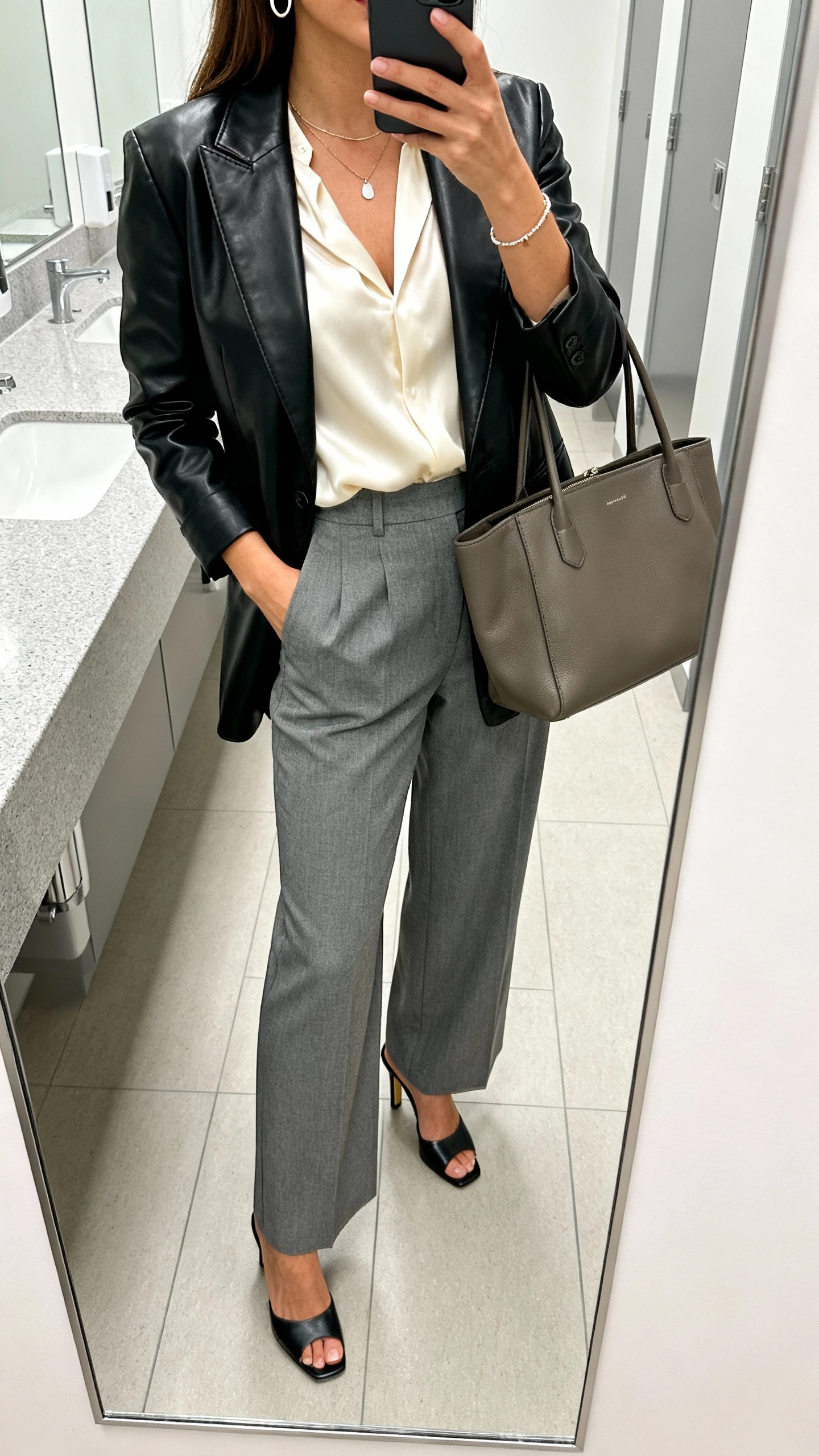 iPhone mirror selfie of a woman in a black leather blazer, cream silk blouse, high-waisted grey wide-leg trousers, heeled mules, structured tote, thin hoops, pendant necklace, and a delicate bracelet; face not visible, office restroom mirror, bright indoor lighting, iPhone photo quality.