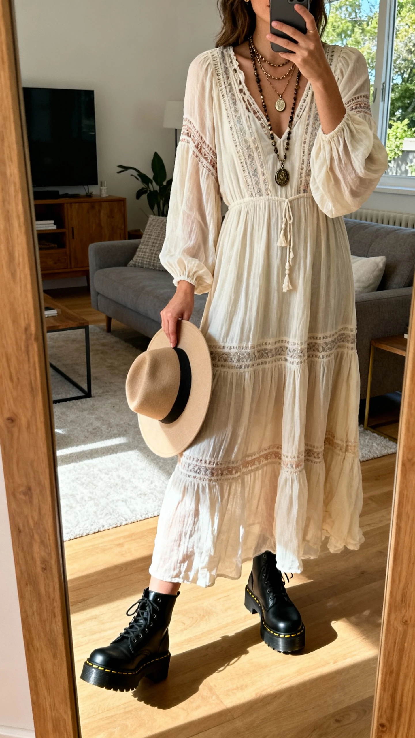 iPhone mirror selfie of a woman in a flowy boho maxi dress with black platform Doc Martens, layered pendant necklaces and a wide-brim hat in hand, face not visible, side angle, living room mirror, natural afternoon light, iPhone photo quality.