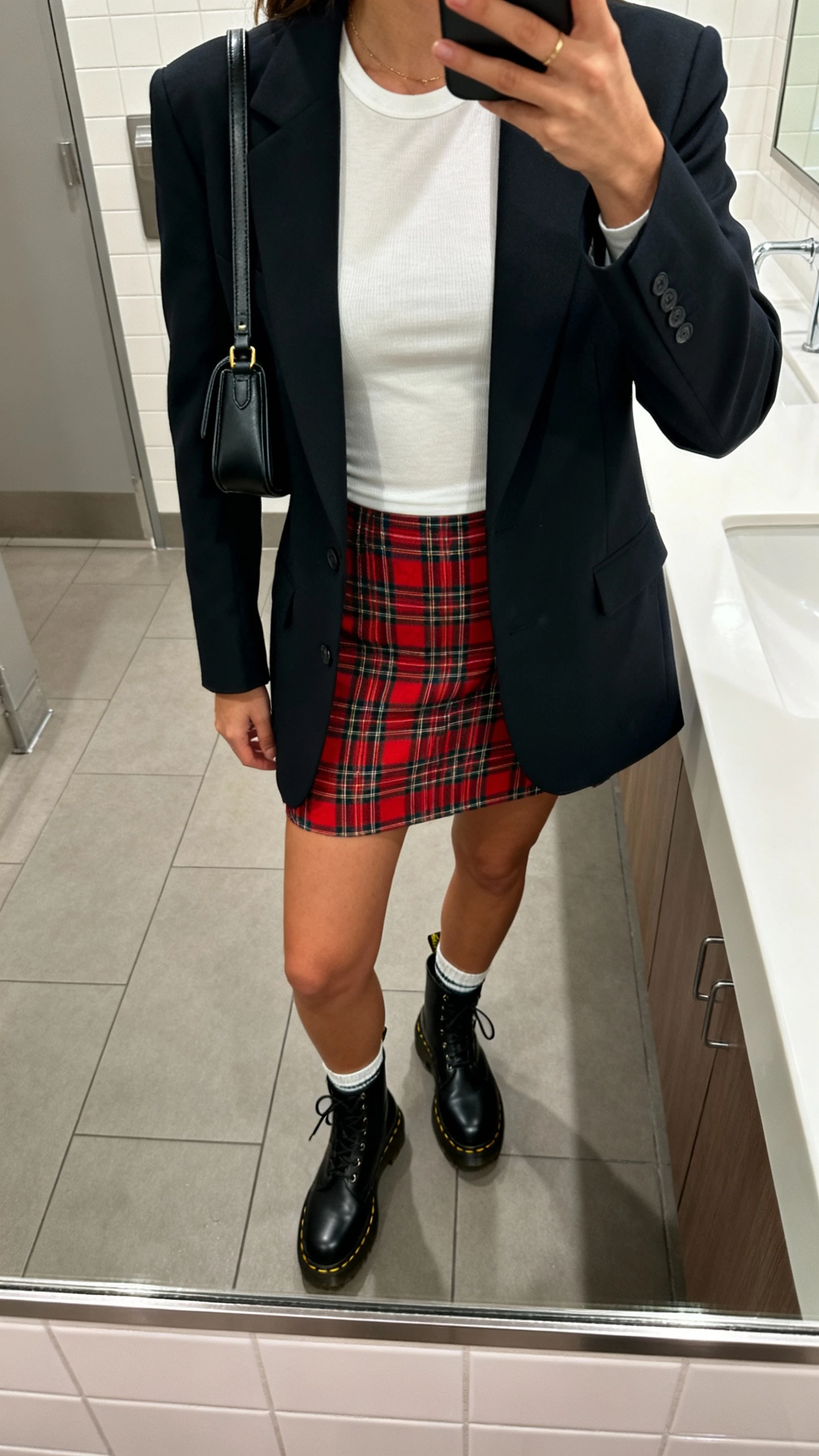 iPhone mirror selfie of a woman in a preppy plaid mini skirt, fitted tee, structured blazer, black Doc Martens and ankle socks, carrying a mini shoulder bag, face not visible, bathroom mirror, overhead indoor lighting, iPhone photo quality.