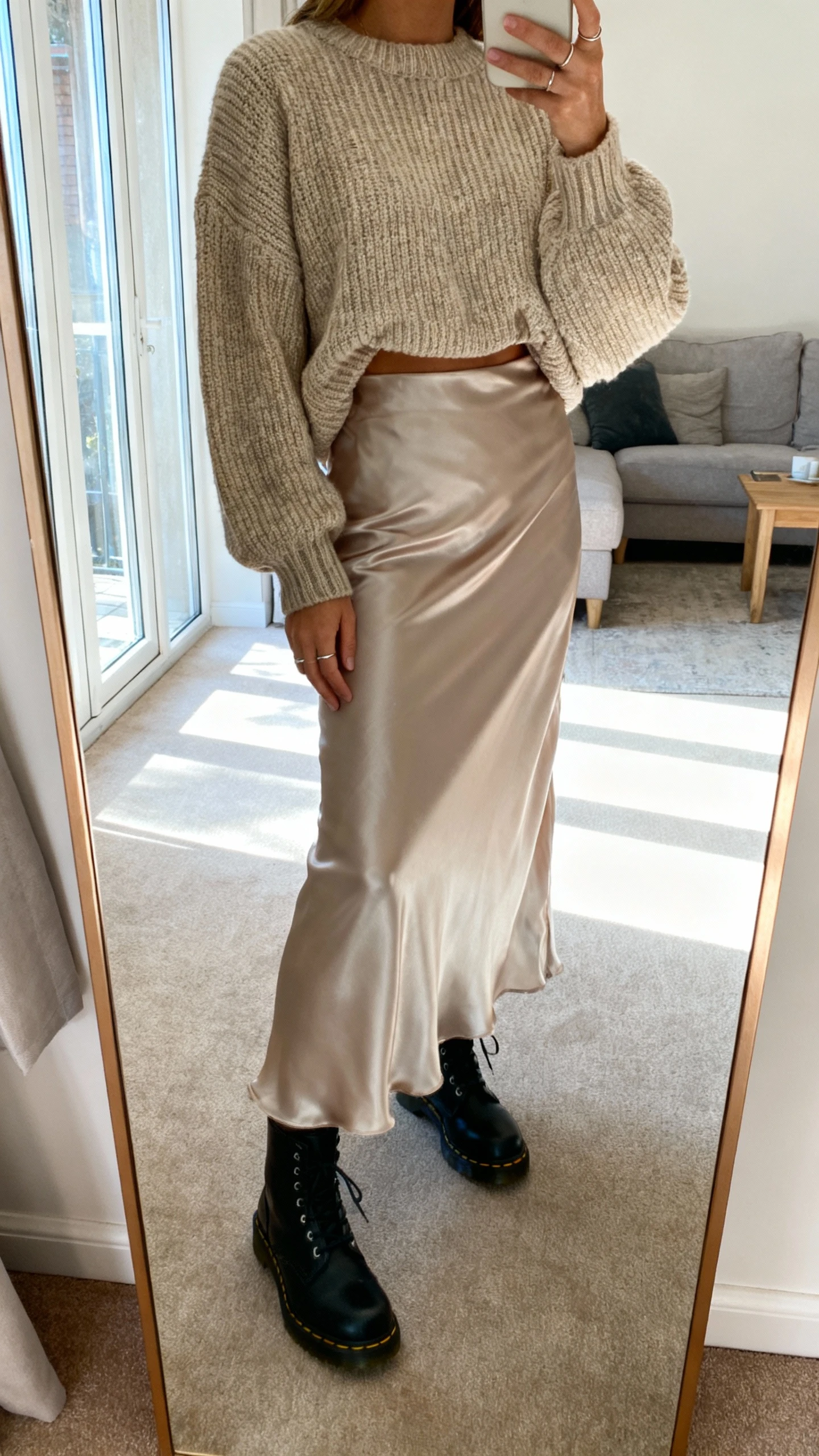 iPhone mirror selfie of a woman in a satiny maxi skirt paired with a chunky knit sweater half-tucked, black Doc Martens, and minimal rings, face not visible, cropped at shoulders, living room full-length mirror, soft window light, iPhone photo quality.