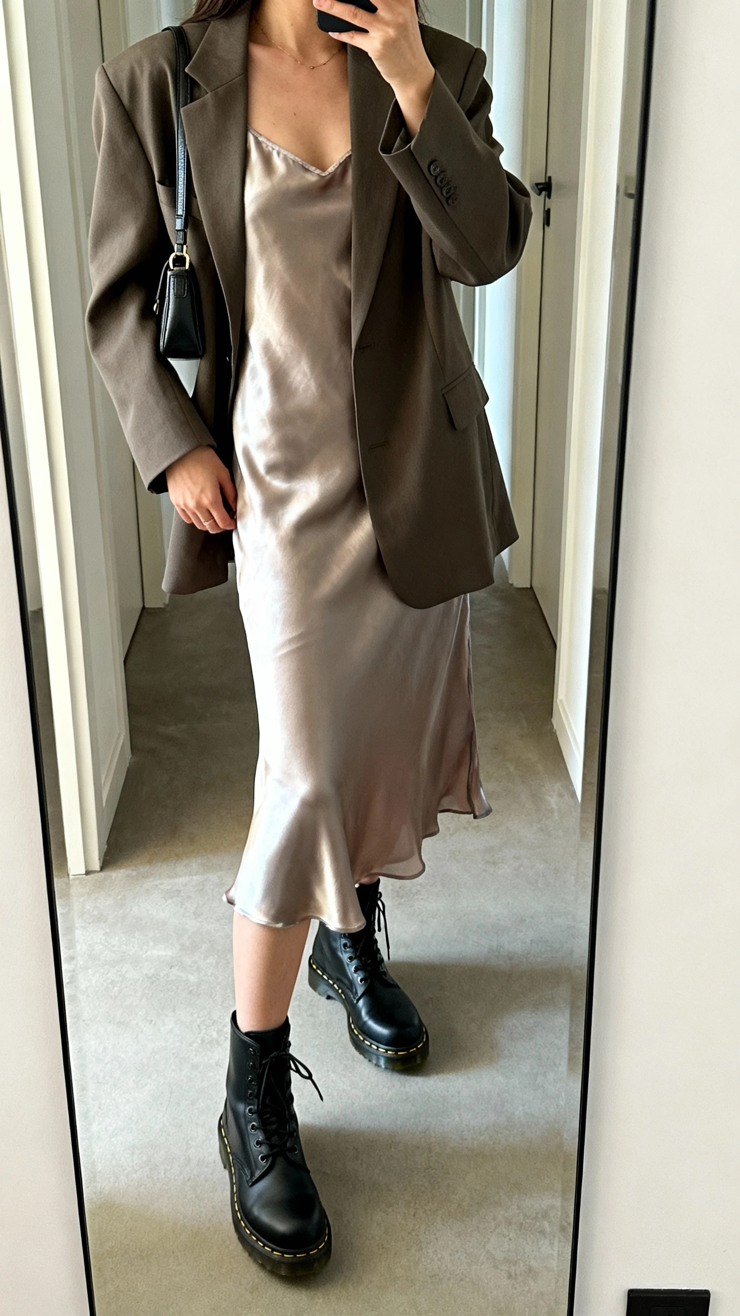 iPhone mirror selfie of a woman in a silky slip dress layered under an oversized blazer with black Doc Martens and a small shoulder bag, face not visible, cropped at shoulders, hallway full-length mirror, indoor daylight, iPhone photo quality.