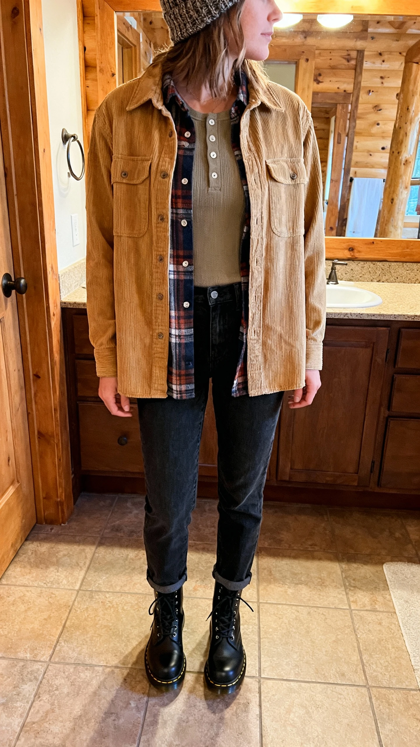 iPhone mirror selfie of a woman in a tan corduroy shirt-jacket layered over a flannel button-up and thermal tee, straight dark jeans, black Doc Martens boots, and a knit beanie, face not visible, cropped at shoulders, cabin-style bathroom mirror with warm indoor light, iPhone photo quality.