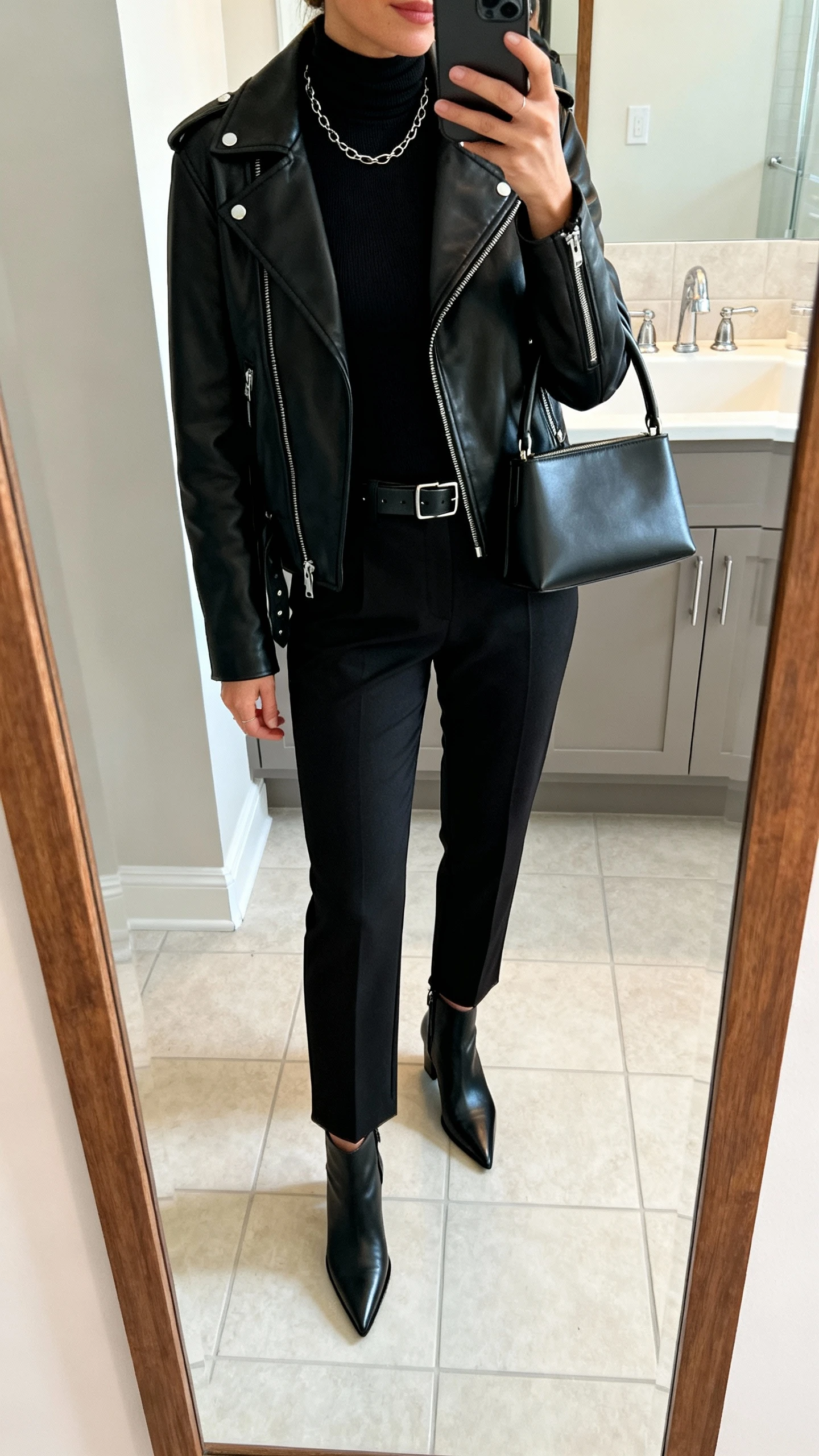 iPhone mirror selfie of a woman in an all-black look: black leather biker jacket, black fitted mock-neck top, black tailored cigarette pants, pointed-toe black ankle boots, minimalist leather belt, thin chain necklace and studs, structured top-handle bag; face not visible, bathroom mirror, soft indoor lighting, iPhone photo quality.