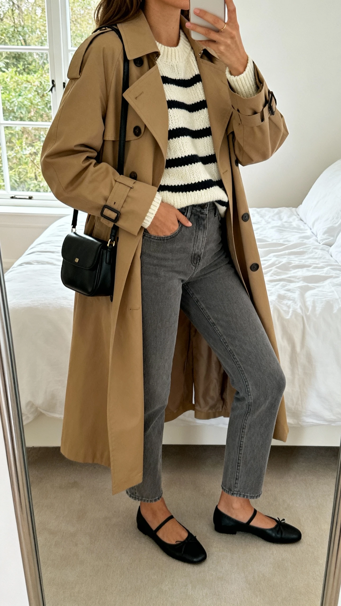iPhone mirror selfie of a woman in grey jeans, a cream-and-black Parisian striped knit, classic tan trench coat, black ballet flats, small black shoulder bag, face not visible, side angle, bedroom mirror, natural window light, iPhone photo quality.