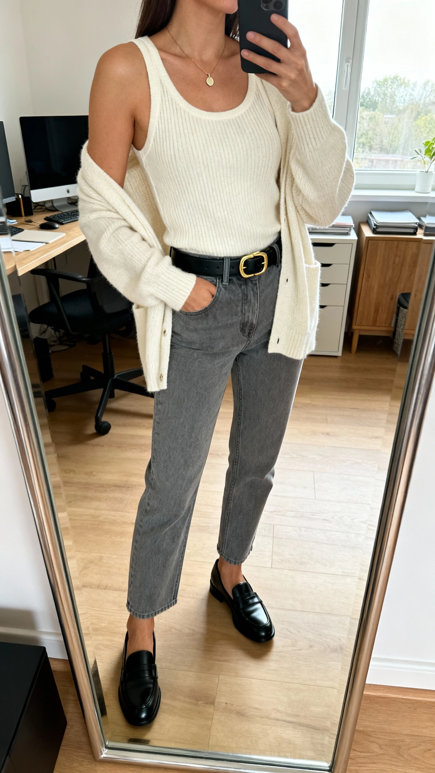 iPhone mirror selfie of a woman in grey jeans, a matching knit cardigan set (tank and cardigan) in cream, black leather loafers, slim black belt with gold buckle, delicate pendant necklace, face not visible, cropped at neck, home office mirror, morning window light, iPhone photo quality.
